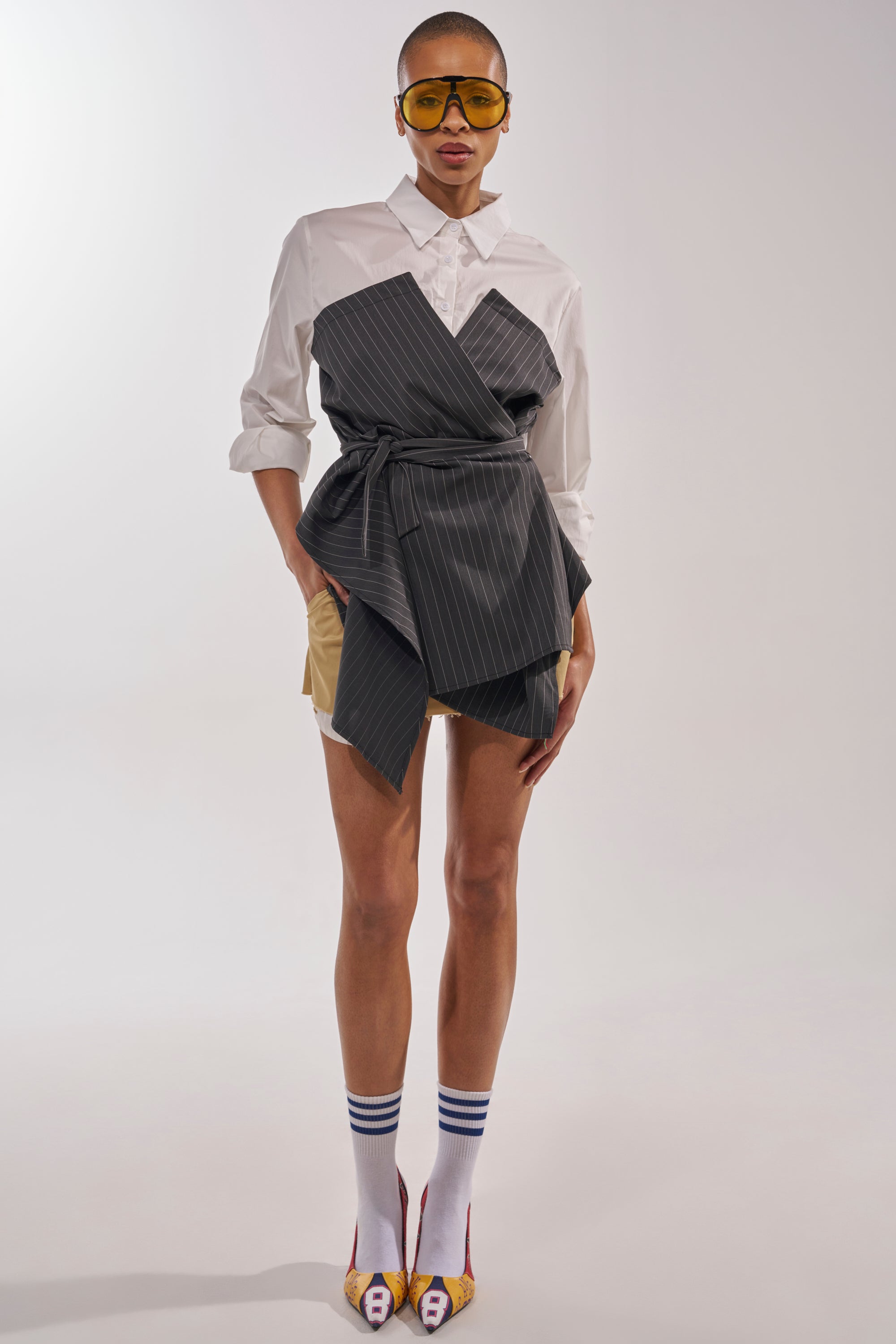 A person faces forward wearing yellow sunglasses, beige shorts, striped white knee-high socks, yellow heeled shoes, a dark wrap top layered over the BUSINESS ONLY BUTTON DOWN pinstripe shirt. The background is plain and light. 7