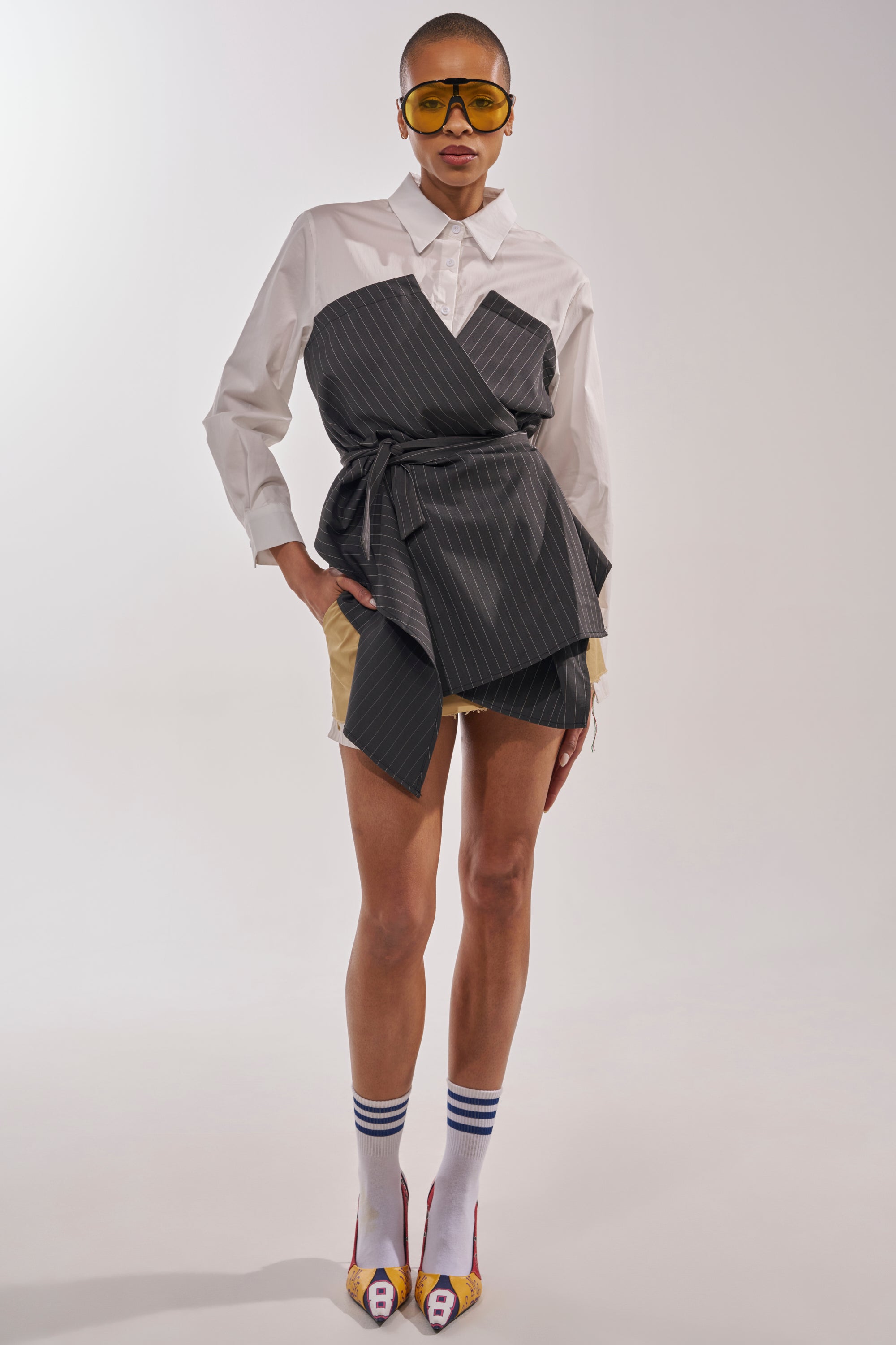 A person stands against a plain background wearing yellow-tinted glasses, beige shorts, white knee-high socks with blue stripes, open-toed heels, and the BUSINESS ONLY BUTTON DOWN shirt for an effortlessly chic office look. 8