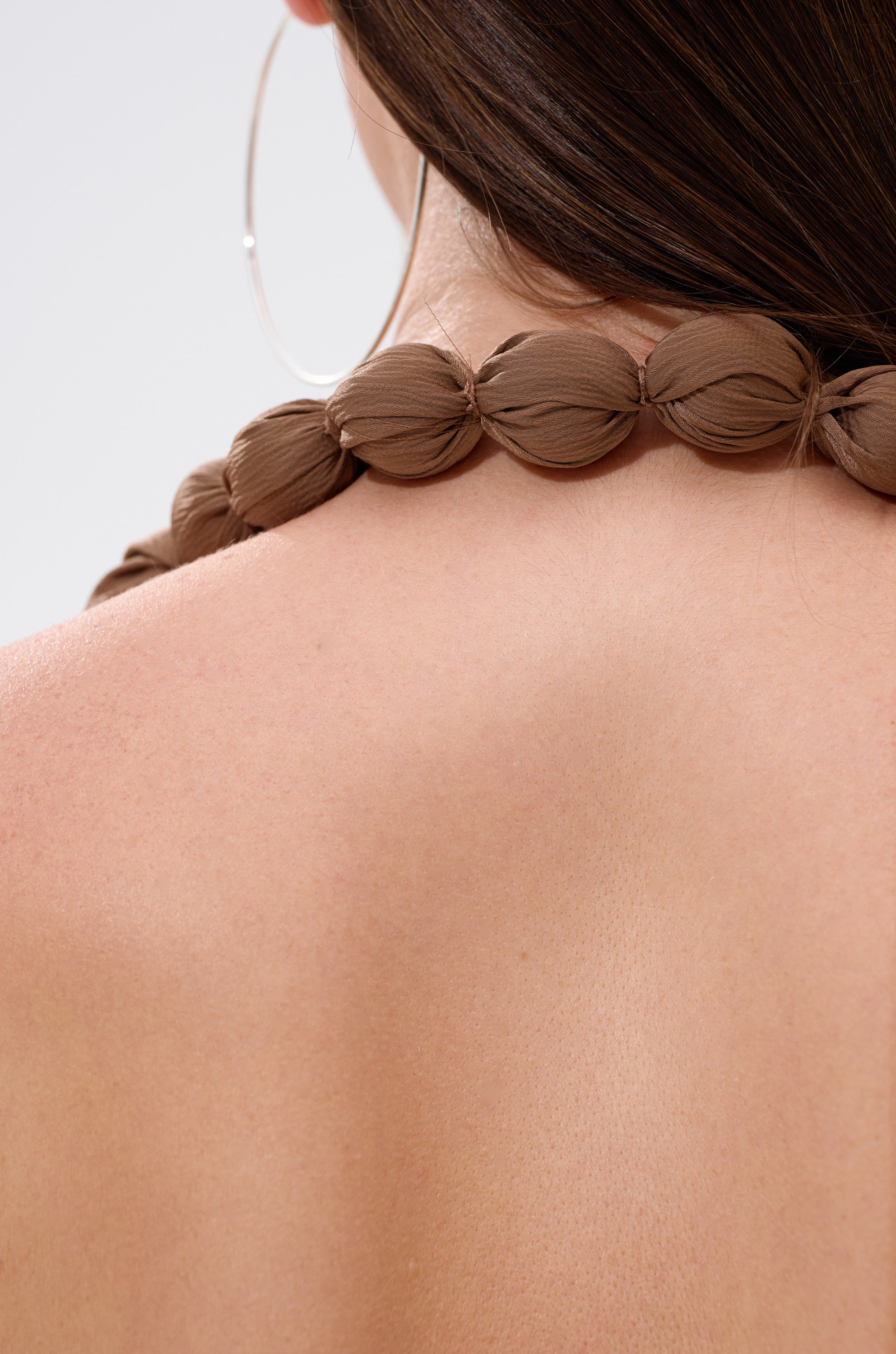 Close-up of a woman wearing the LEXI SATIN COWL NECK BLOUSE, her bare back and shoulder highlighted alongside a large hoop earring and brown beaded necklace. Her straight brown hair complements the blouse’s elegant open-back design.