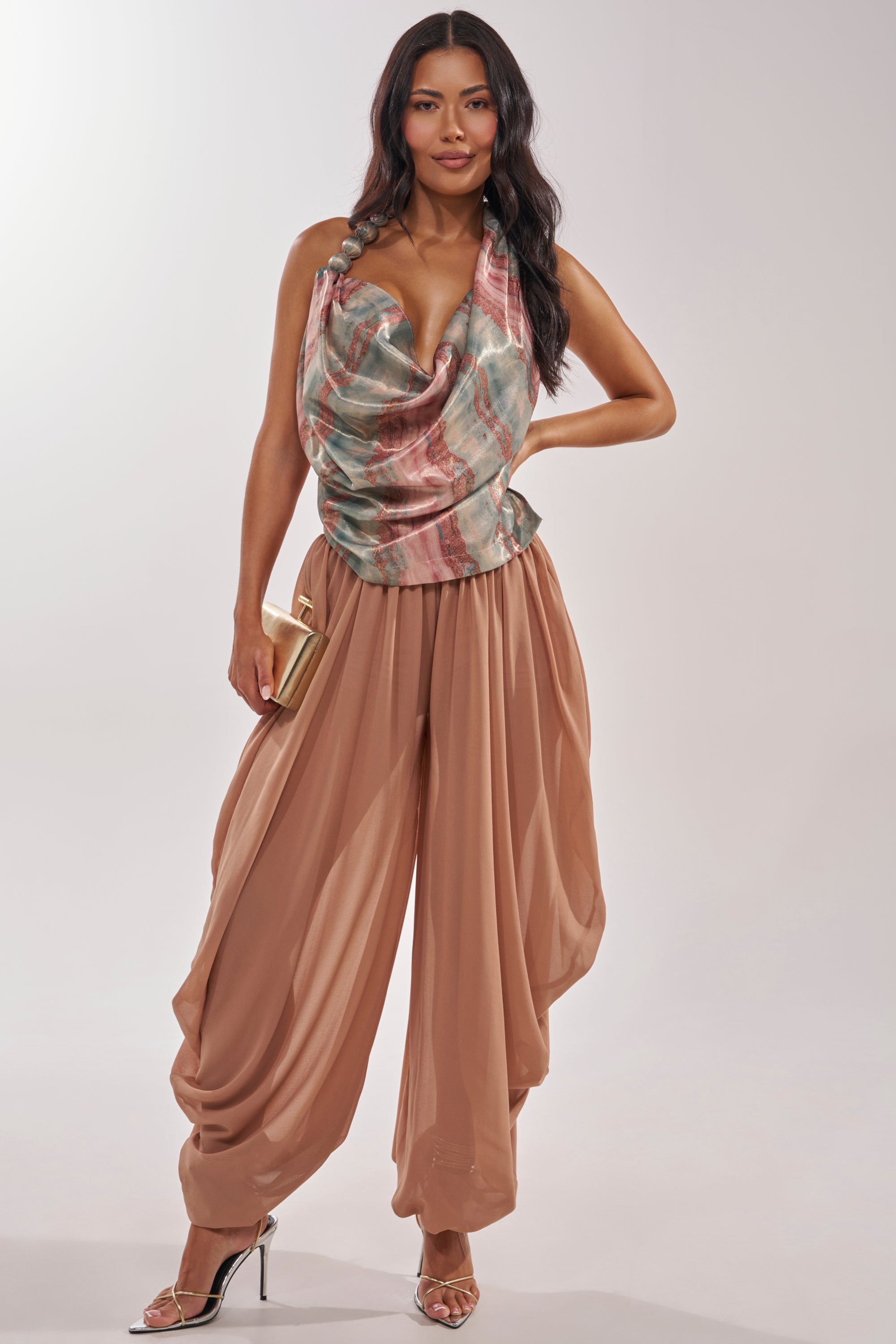 A woman poses in loose tan harem pants, gold heels, and a gold clutch, paired with the vibrant FEEL THE VIBES BLOUSE—a bubble strap halter top with a cowl neckline—creating an eye-catching festival look against a plain light background. 6