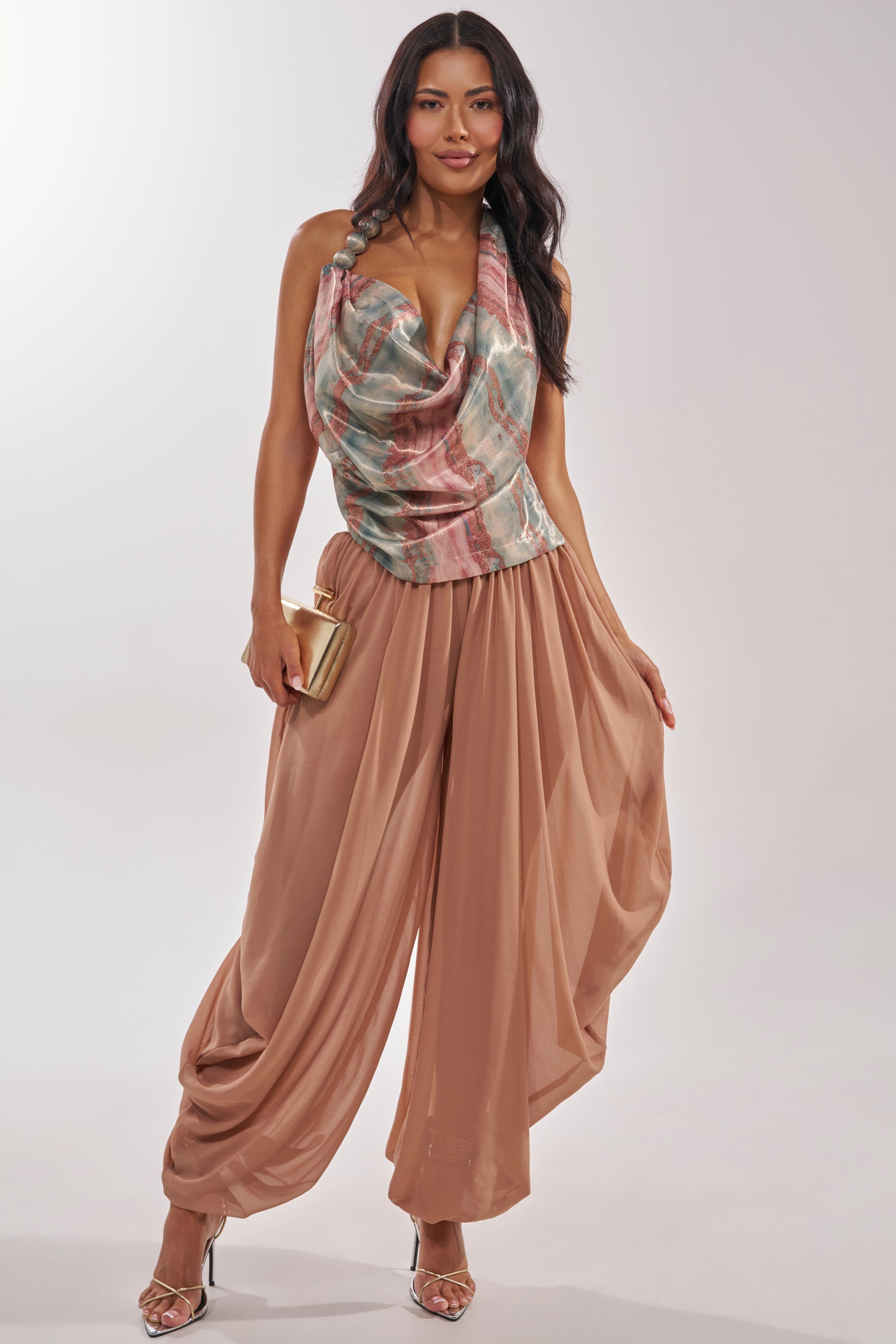 A woman poses in the FEEL THE VIBES BLOUSE—a sleeveless, draped multicolor halter top—paired with wide taupe pants, gold clutch, and high-heeled sandals for a chic festival look against a light background. 7