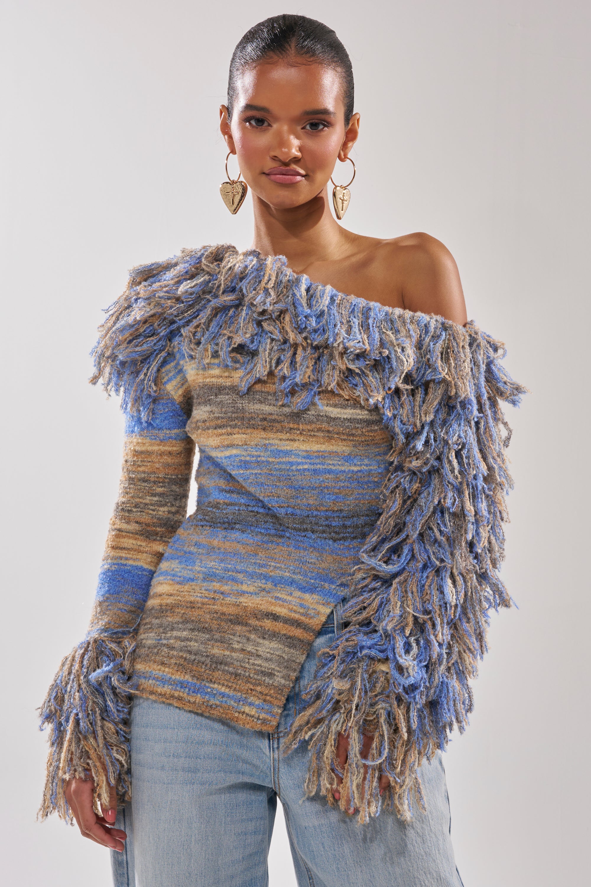 A woman stands confidently against a plain light background, wearing the FRINGE FOR DAYS SWEATER—a multicolor striped fringe sweater with one off-the-shoulder sleeve—paired with light blue jeans.