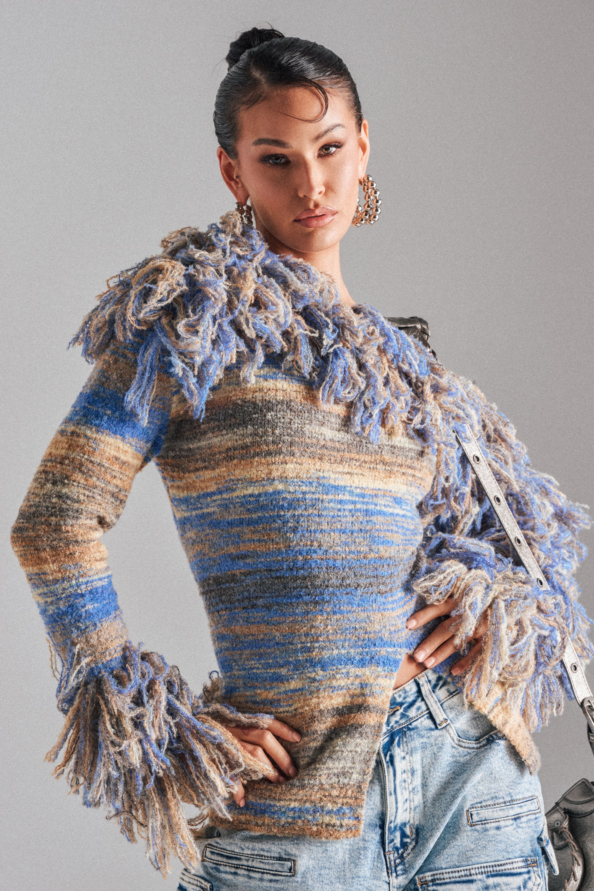 A woman poses confidently in the FRINGE FOR DAYS SWEATER, a multicolor striped knit with chunky fringed shoulders and cuffs, styled with light blue jeans against a plain background.