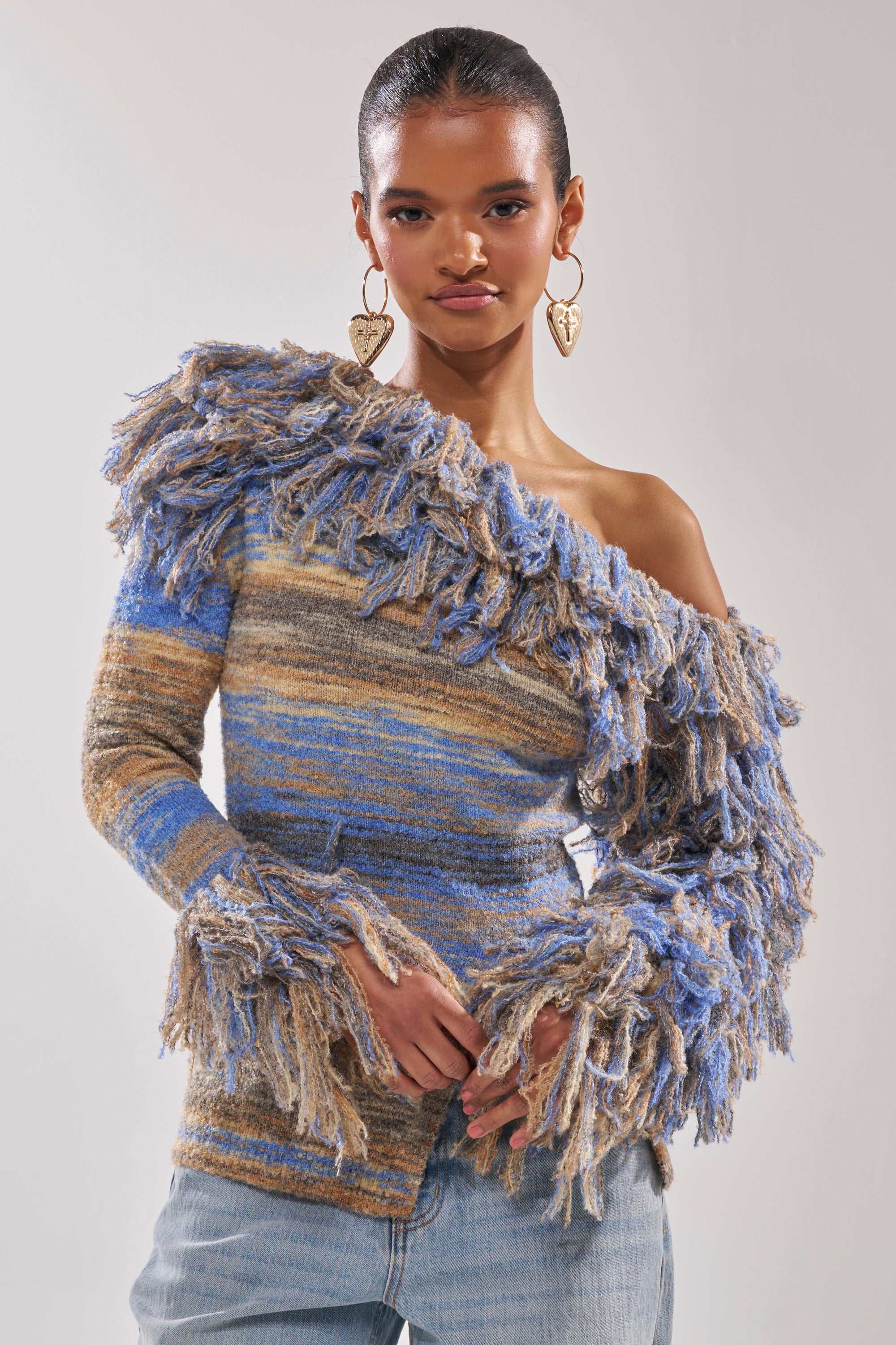 A woman with slicked-back hair wears heart-shaped earrings and the FRINGE FOR DAYS SWEATER—an off-the-shoulder, multicolor striped knit in blue, brown, and beige—paired with light blue jeans against a plain background.