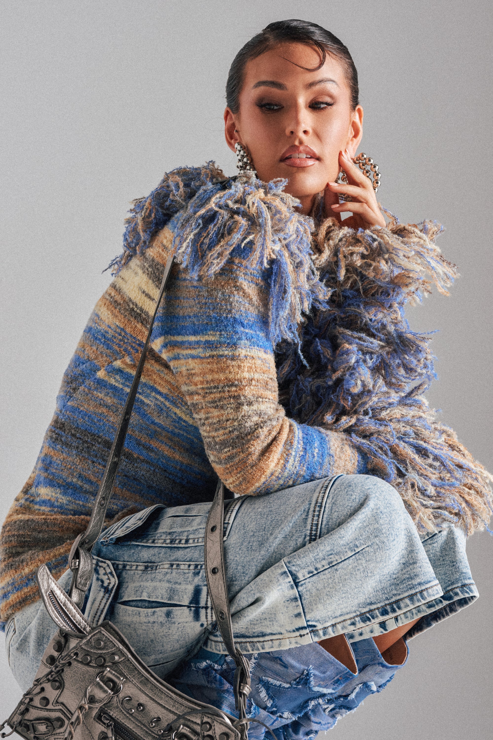 A woman poses confidently in the FRINGE FOR DAYS SWEATER featuring multicolor stripes with blue and brown tones, paired with distressed jeans, a metallic silver handbag, slicked-back hair, and large earrings.