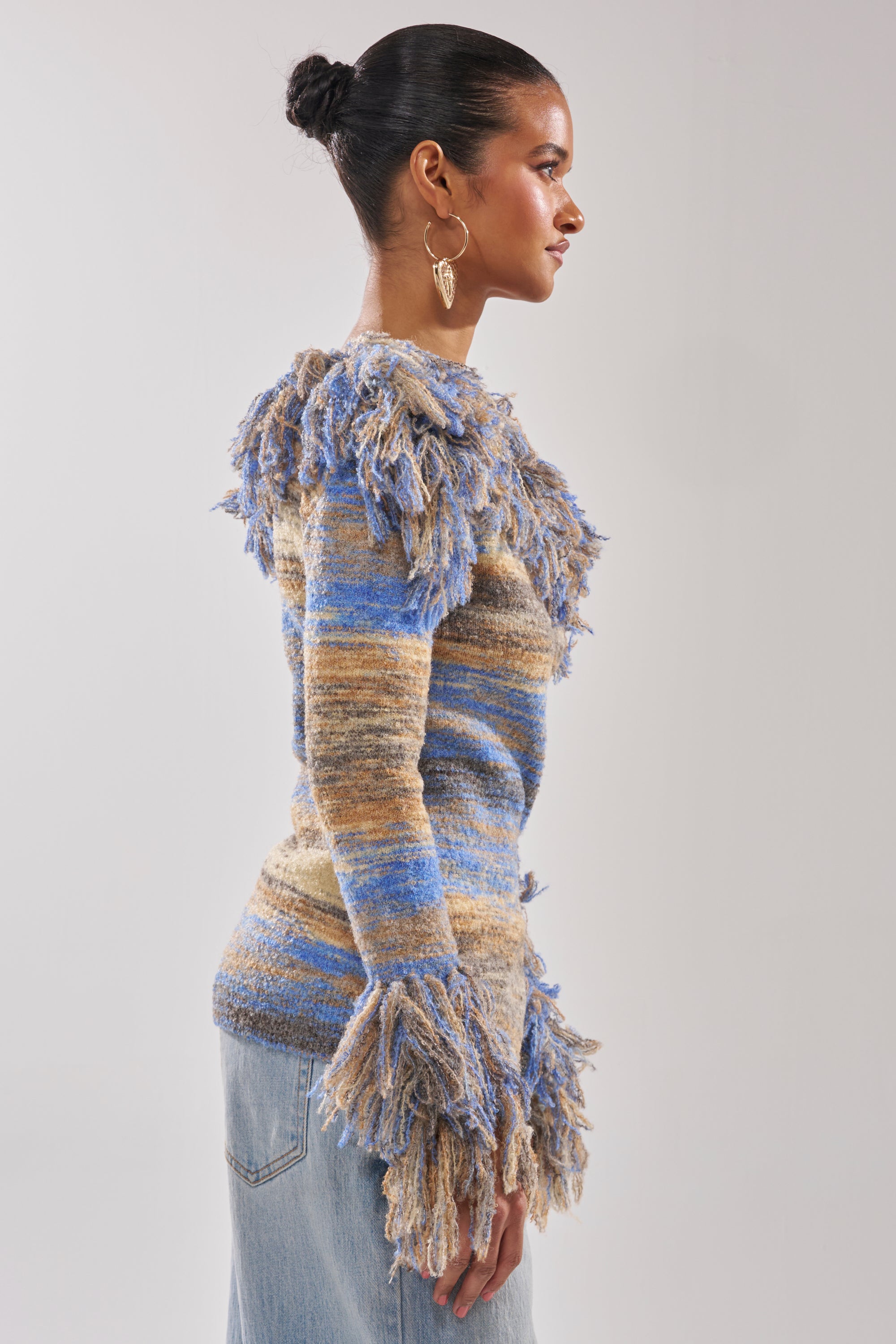 A woman in profile wears the FRINGE FOR DAYS SWEATER, featuring multicolor stripes in blue, beige, and brown. She styles it with her dark hair in a bun, large hoop earrings, and light blue jeans for a chic, effortless vibe.