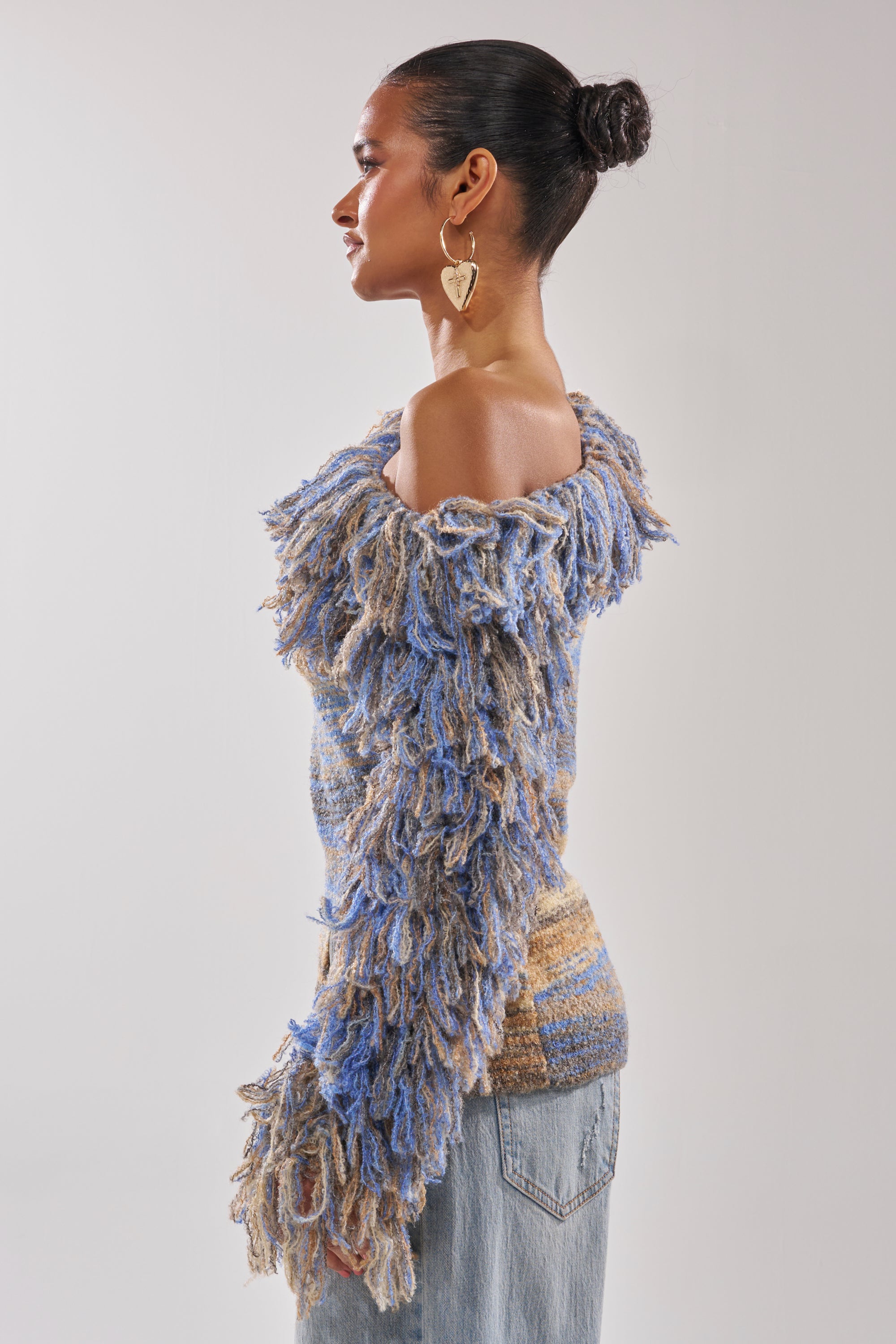 A woman stands in profile against a plain background, wearing the FRINGE FOR DAYS SWEATER in blue and beige with textured knit fringe details, paired with light denim jeans. Her hair is styled in a bun and she wears large heart-shaped earrings.