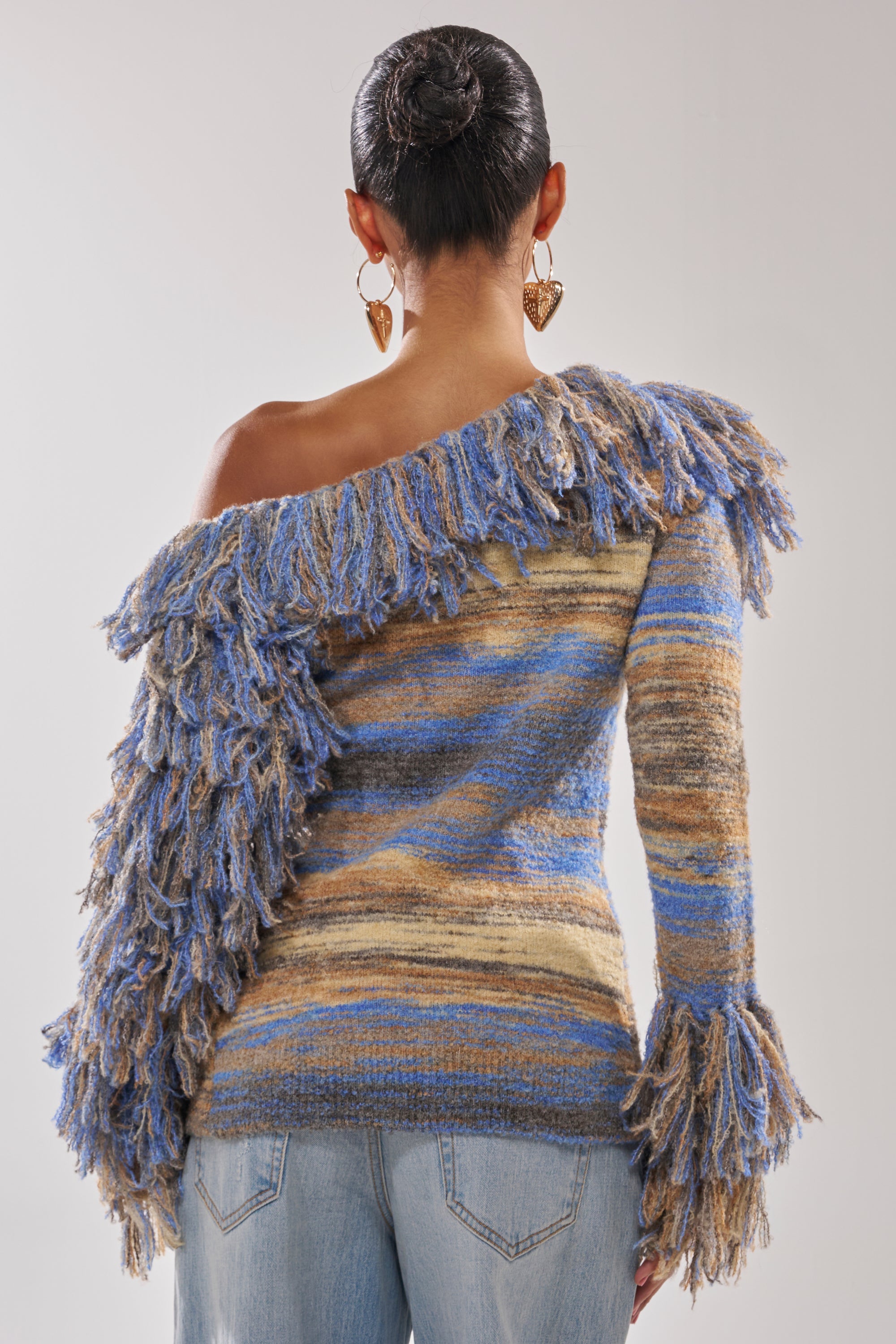 A person faces away, wearing the FRINGE FOR DAYS SWEATER—a multicolor knit, off-the-shoulder style with blue, beige, and brown stripes plus fringe—paired with light blue jeans. Their hair is styled in a neat bun.