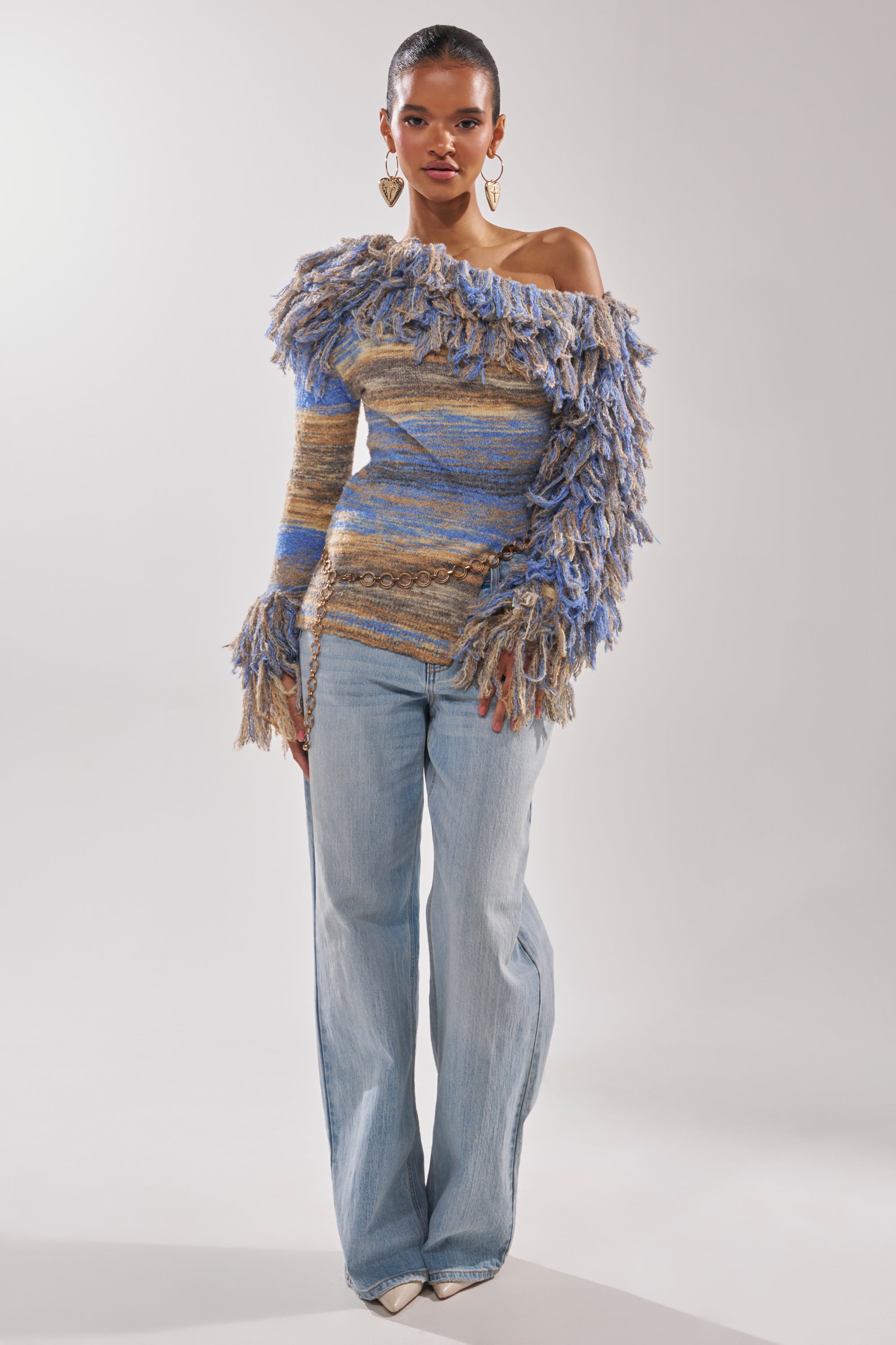 A woman stands against a plain background wearing wide-leg light blue jeans and the FRINGE FOR DAYS SWEATER—an off-the-shoulder knit with blue and beige stripes plus fringed detail on one sleeve. She accessorizes with gold heart-shaped earrings.