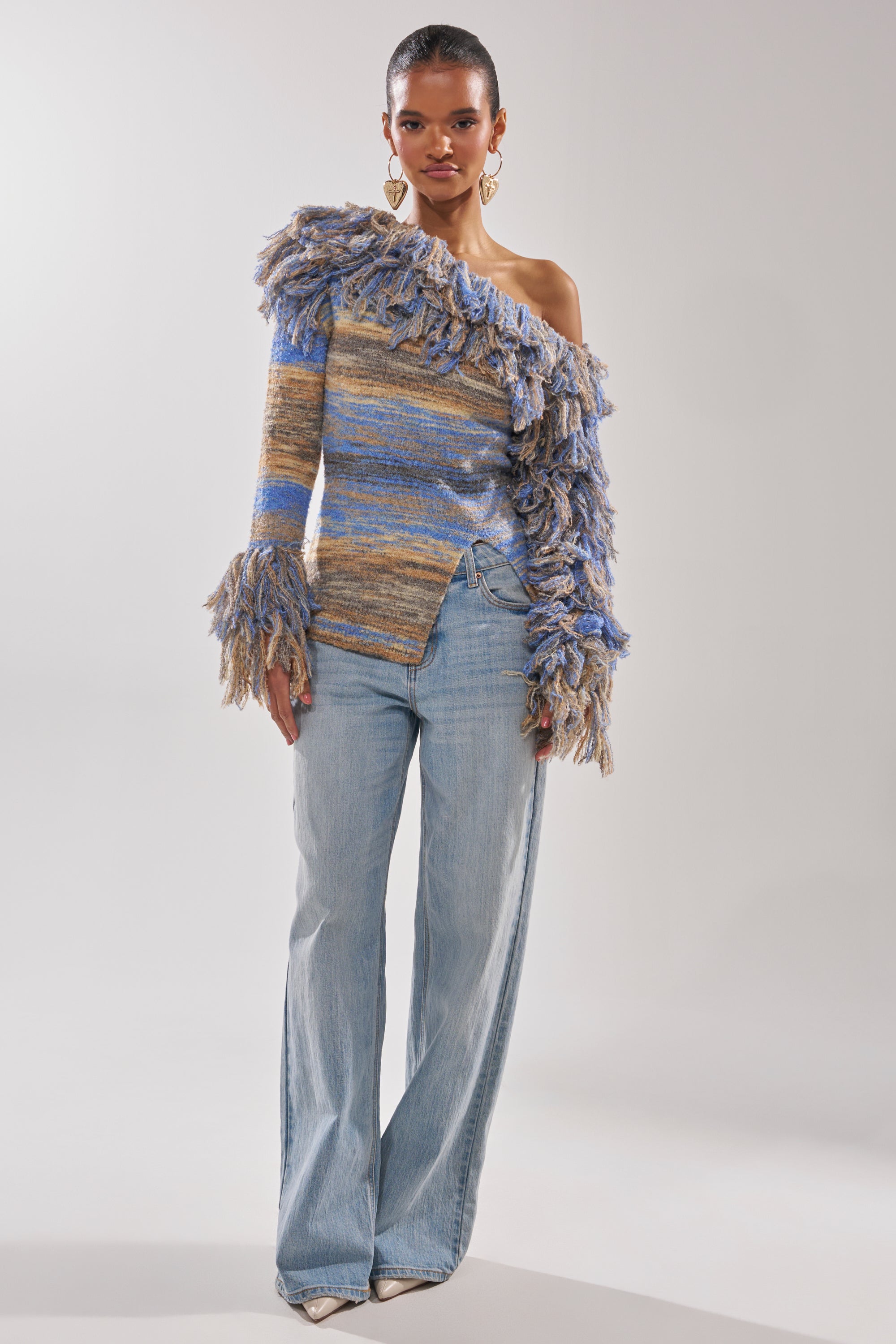 A woman poses in the FRINGE FOR DAYS SWEATER, a multicolor striped off-the-shoulder knit with fringed ruffles, paired with light blue wide-legged jeans, gold heart earrings, and white pointed shoes against a plain background.
