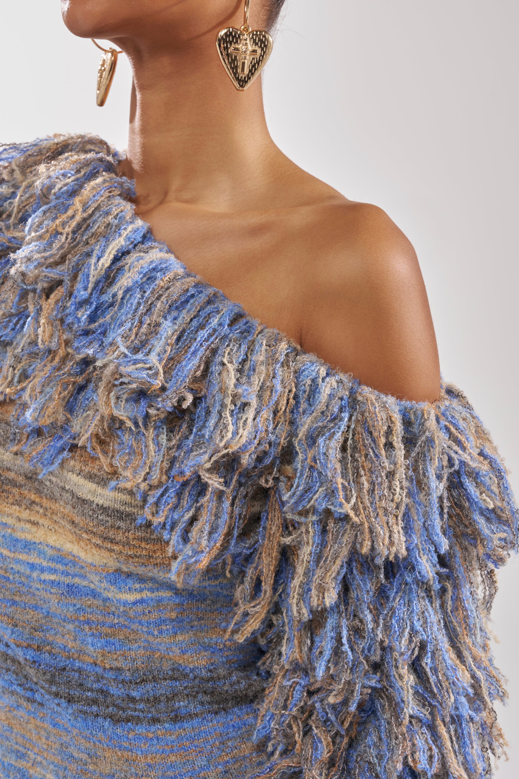 Wearing the FRINGE FOR DAYS SWEATER—a textured, multicolor striped off-the-shoulder knit with blue, beige, and brown fringe—paired with large heart-shaped earrings.