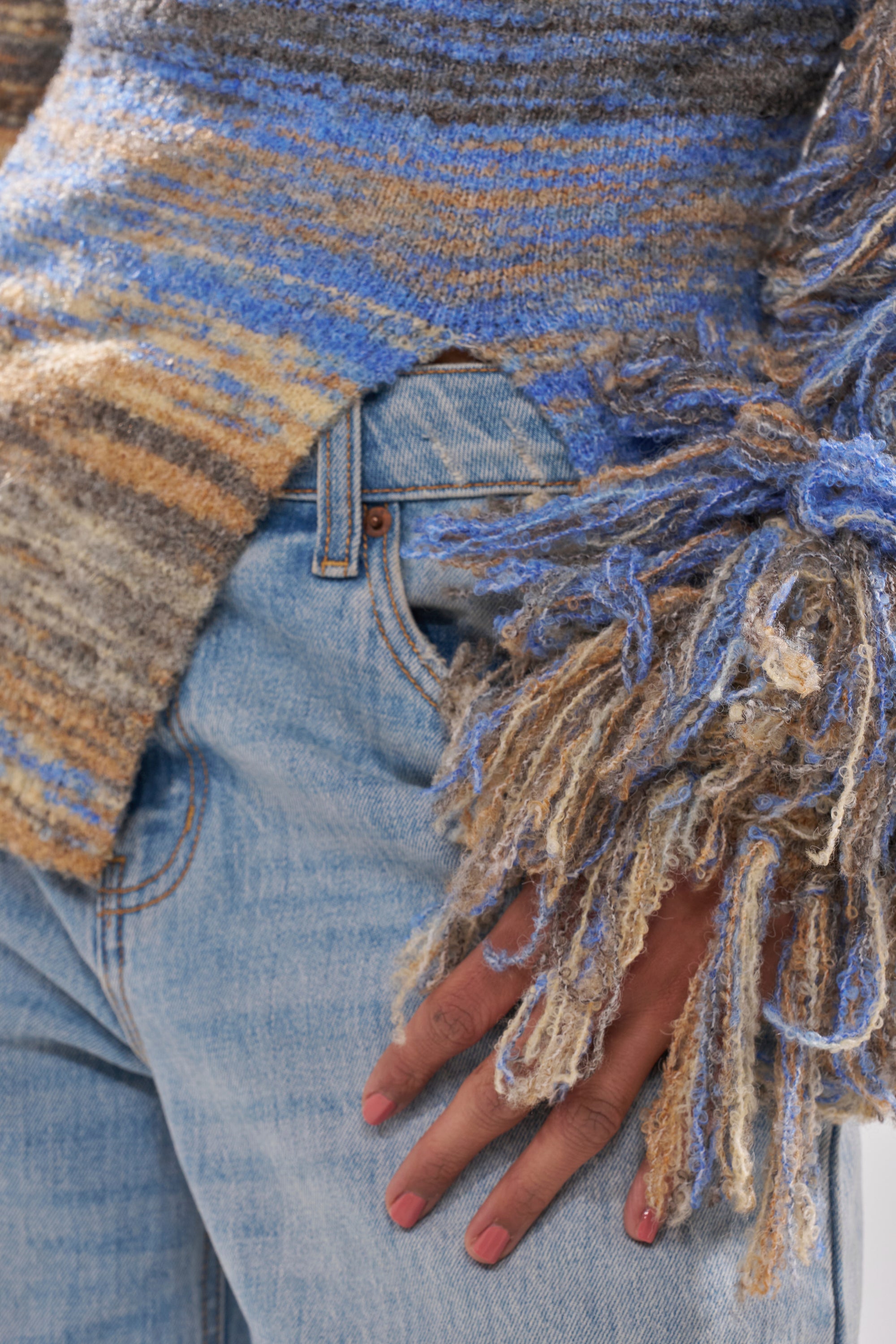 A close-up of a person wearing light blue jeans and the FRINGE FOR DAYS SWEATER, a multicolor striped piece in blue, brown, and beige with long, thick fringe details on the sleeve cuffs.