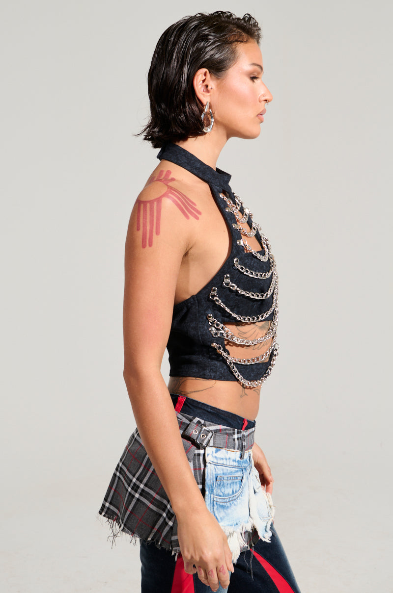 A person with short dark hair stands in profile, exuding skeleton vibes in a TINY LIL GIRL HALTER TOP WITH CHAIN, distressed denim pants, and a red and gray plaid accessory. They showcase a handprint tattoo on their shoulder and large hoop earrings. 1