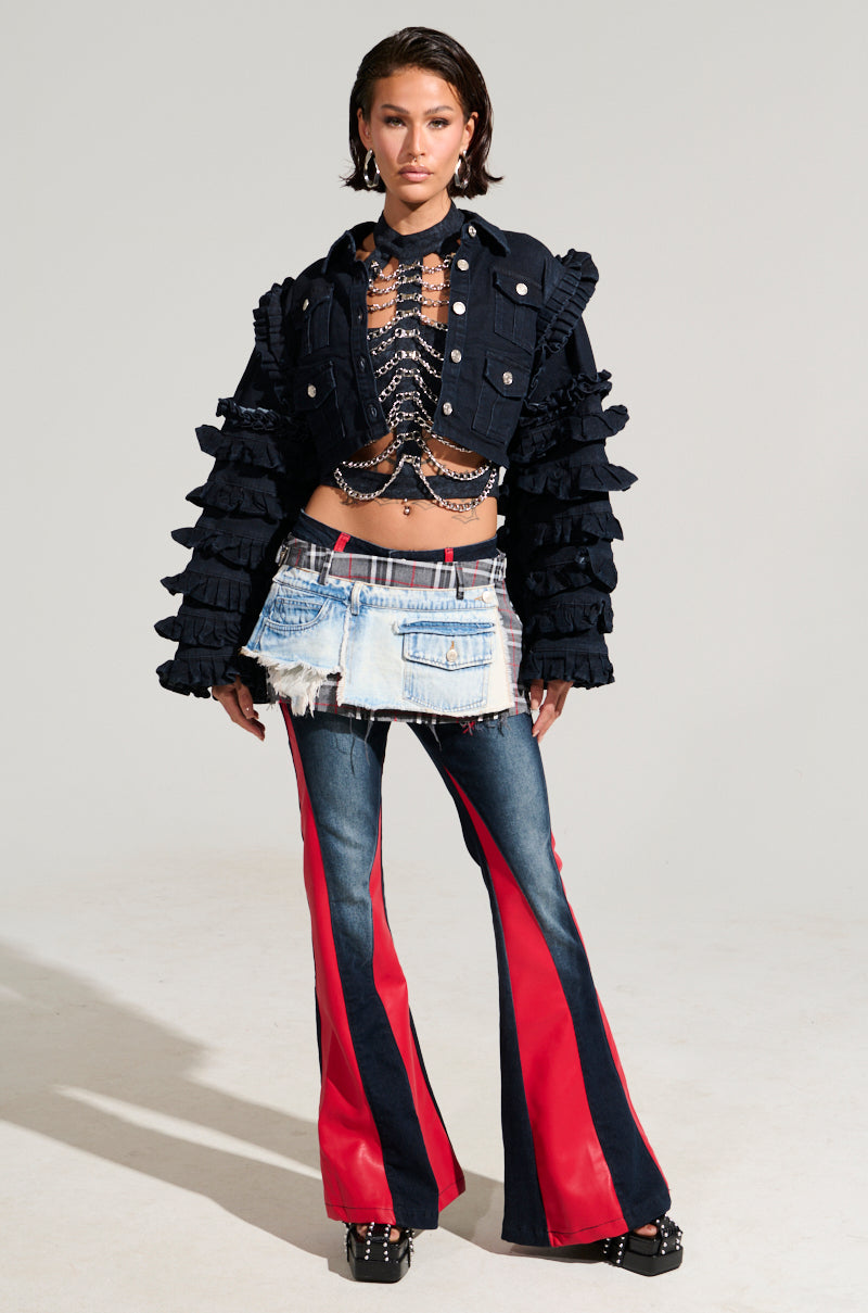 A person poses confidently in a fashionable outfit comprising the TINY LIL GIRL HALTER TOP WITH CHAIN, paired with a black ruffled jacket featuring silver chain details, a distressed denim mini skirt layered over red and blue flared jeans, and black studded shoes. The ensemble exudes subtle skeleton vibes against a plain gray background. 5