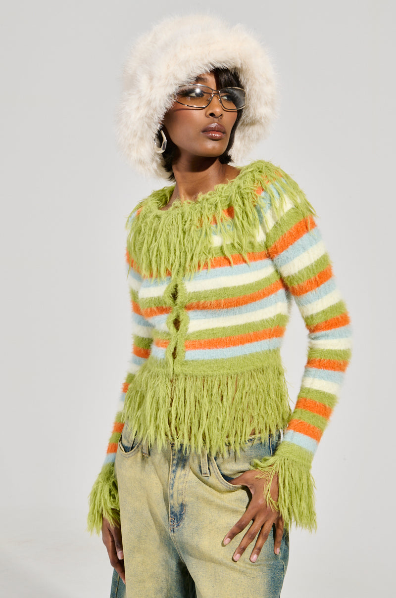 Dressed in the COZY AND CUTE SWEATER, a person wearing a fluffy white hat, oversized sunglasses, and hoop earrings stands confidently. Their fuzzy sweater features multicolored stripes with green fringe and pairs effortlessly with faded jeans. A simple gray backdrop highlights their vibrant style.