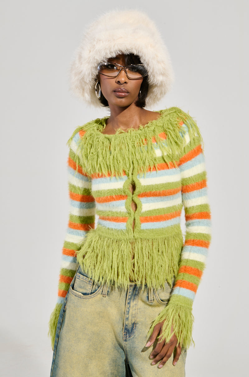 A person stands confidently, sporting a fluffy white hat and sunglasses. They're dressed in the COZY AND CUTE SWEATER, featuring a colorful pattern with green, orange, and white stripes, paired with light-colored jeans against a simple, neutral background.