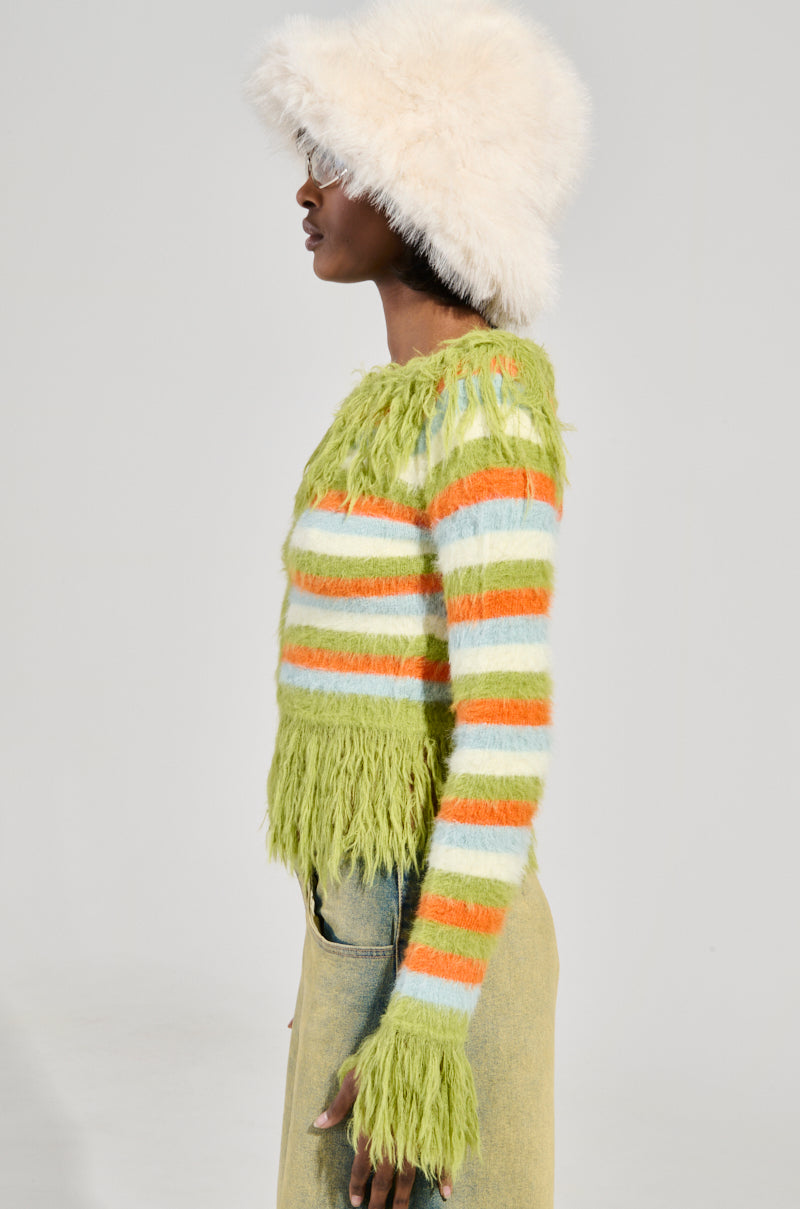 A person in profile wearing a fluffy hat and the COZY AND CUTE SWEATER, which features a cozy, cute striped design in green, orange, and white. The off-the-shoulder sweater highlights long sleeves and a shaggy texture. They pair it with light-colored pants against a plain background.