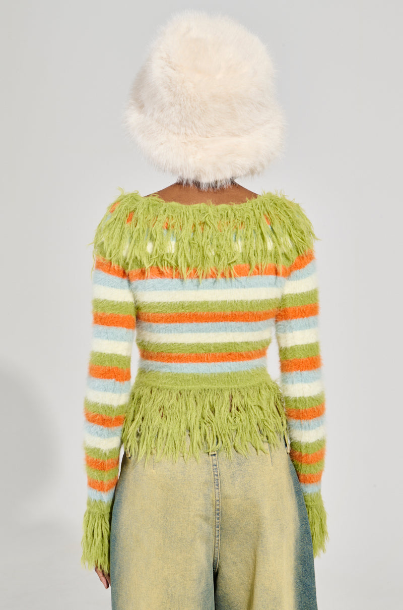A person faces away, wearing a fluffy white hat and the COZY AND CUTE SWEATER—a vibrant green, orange, and white striped cardigan adorned with fringe details. They expertly pair it with faded jeans. The plain, light-colored background enhances the playful charm of their outfit.