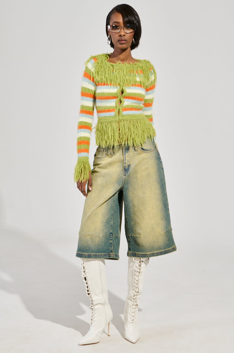 A person wearing the COZY AND CUTE SWEATER in green and orange stripes, paired with wide-legged denim shorts and knee-high white lace-up boots, stands against a plain background. They are sporting sunglasses and exude confidence in their pose.