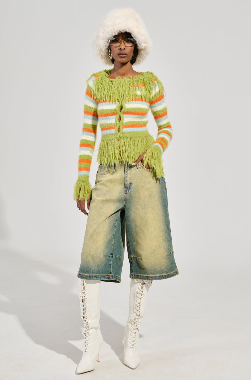 Dressed in a COZY AND CUTE SWEATER, featuring fluffy stripes in green and orange, paired with denim shorts, white knee-high lace-up boots, sunglasses, and a white furry hat, a person poses against a plain background.