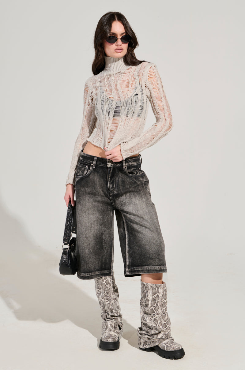 A person wearing dark sunglasses, a SEXY LOOSE WEAVE HIGH NECK SWEATER IN GREY, faded knee-length shorts, and knee-high snake print boots poses confidently while holding a black bag.