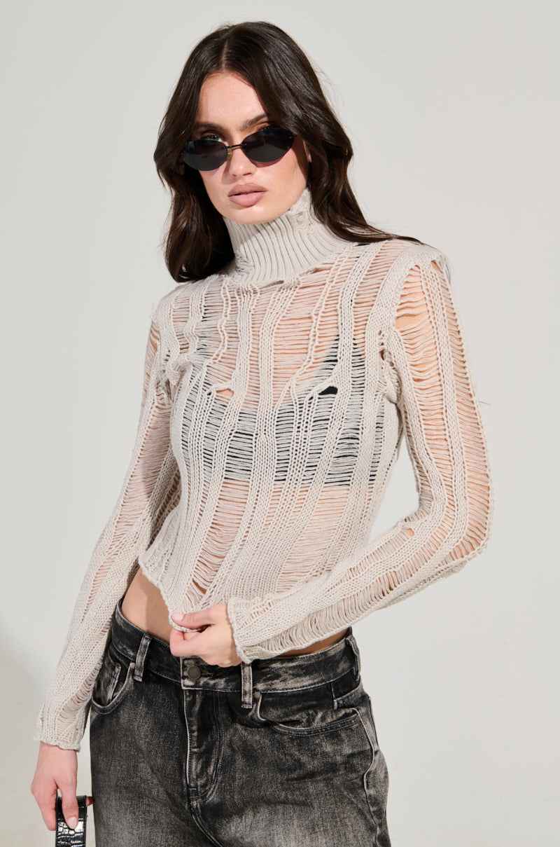 A person with long hair is wearing sunglasses and the SEXY LOOSE WEAVE HIGH NECK SWEATER IN GREY, which boasts a sheer, high-neck design with vertical, distressed paneled detailing, paired perfectly with dark, faded jeans. The plain background amplifies the striking outfit.