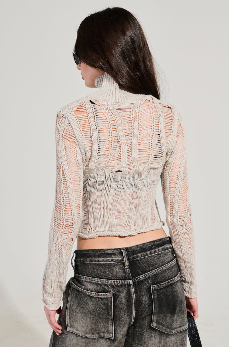 A person with long dark hair and sunglasses is facing away from the camera, dressed in a sexy loose weave high neck sweater in grey with distressed paneled detailing paired with black jeans. They are holding a black handbag against a plain, light-colored background.