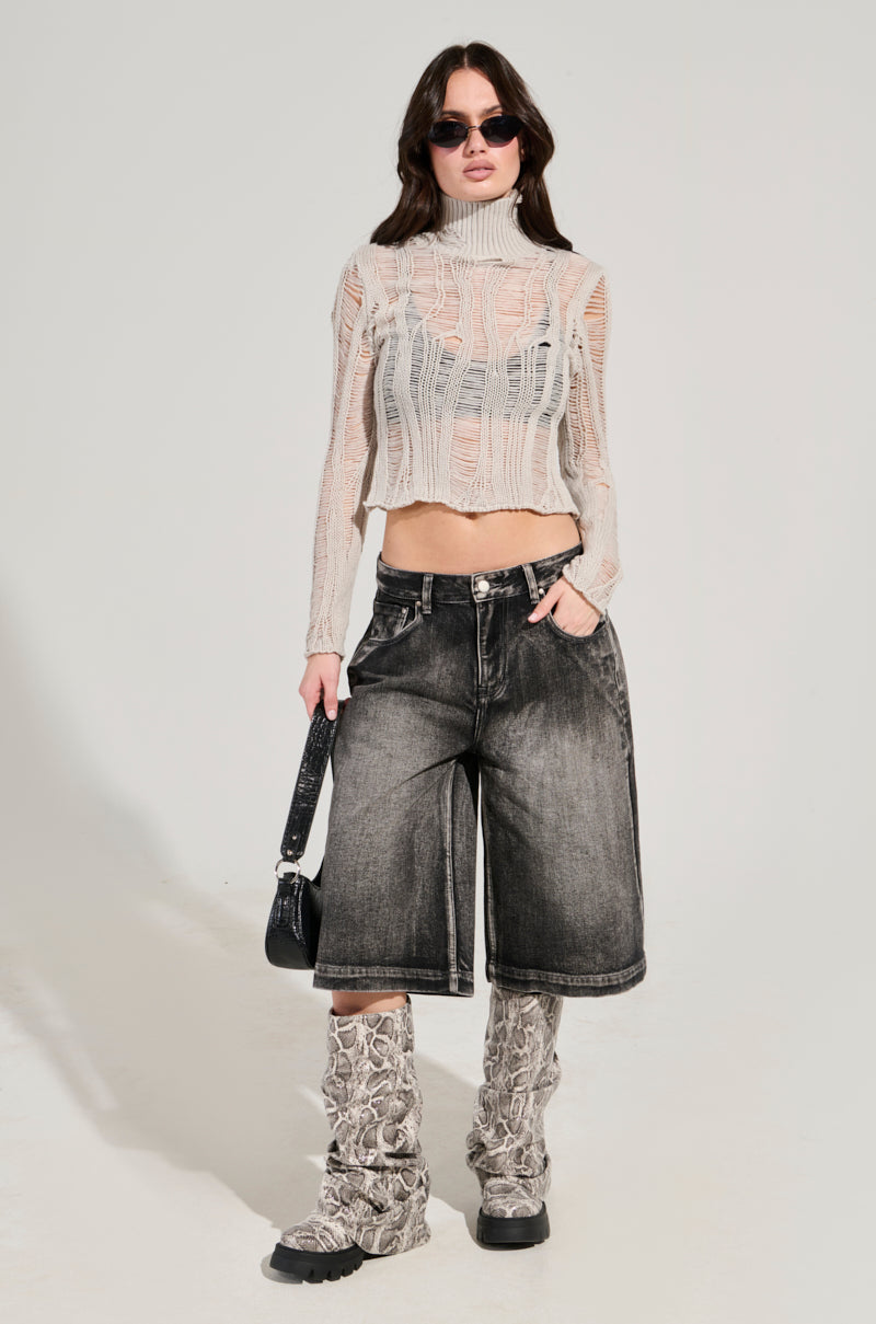 A person wearing sunglasses, a SEXY LOOSE WEAVE HIGH NECK SWEATER IN GREY, denim culottes, and patterned knee-high boots stands against a plain background, holding a small black bag by their side. They exhibit a confident style.