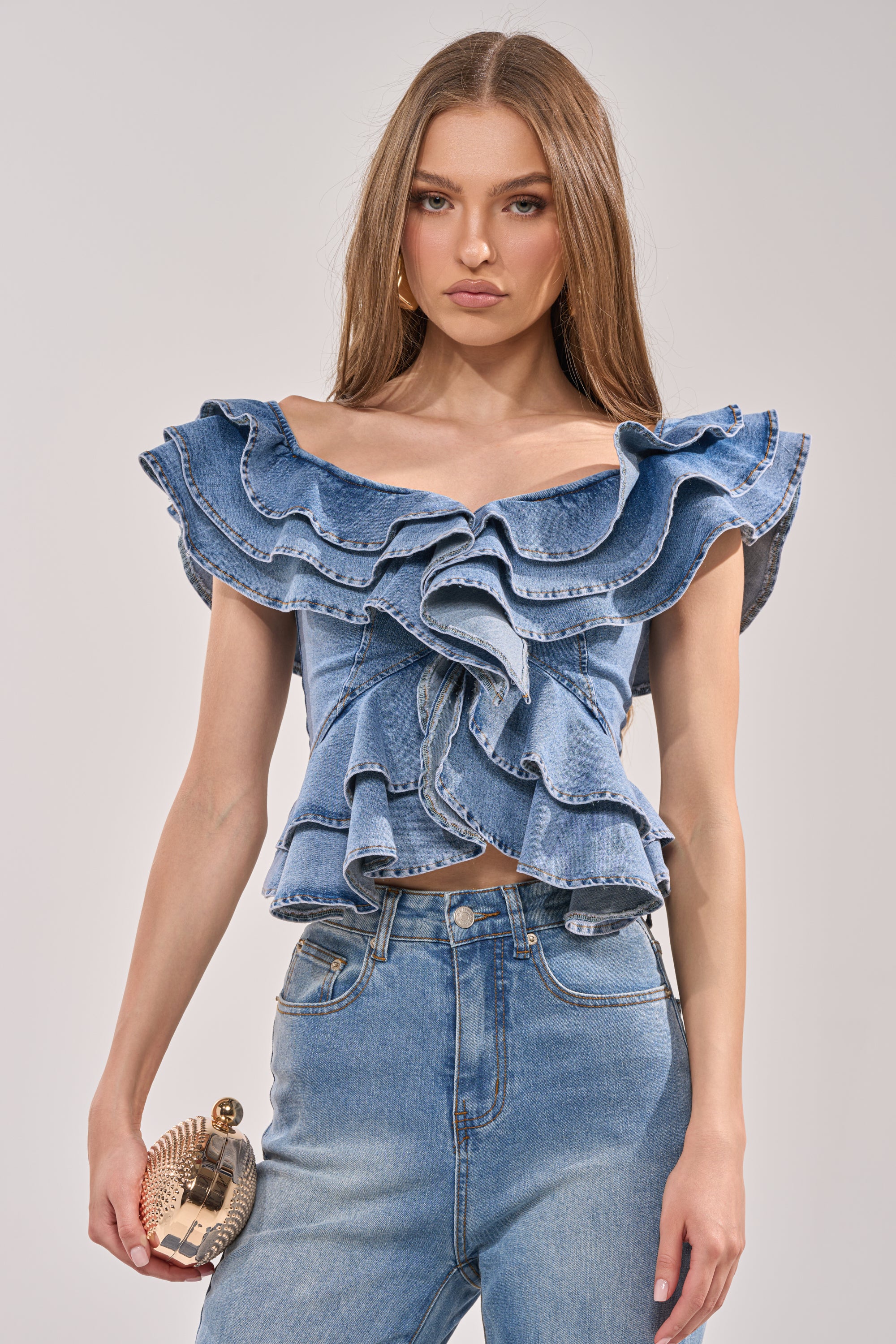 A woman poses against a plain light background wearing the DREAMING BIG DENIM RUFFLE TOP and high-waisted jeans, holding a textured clutch purse. 0
