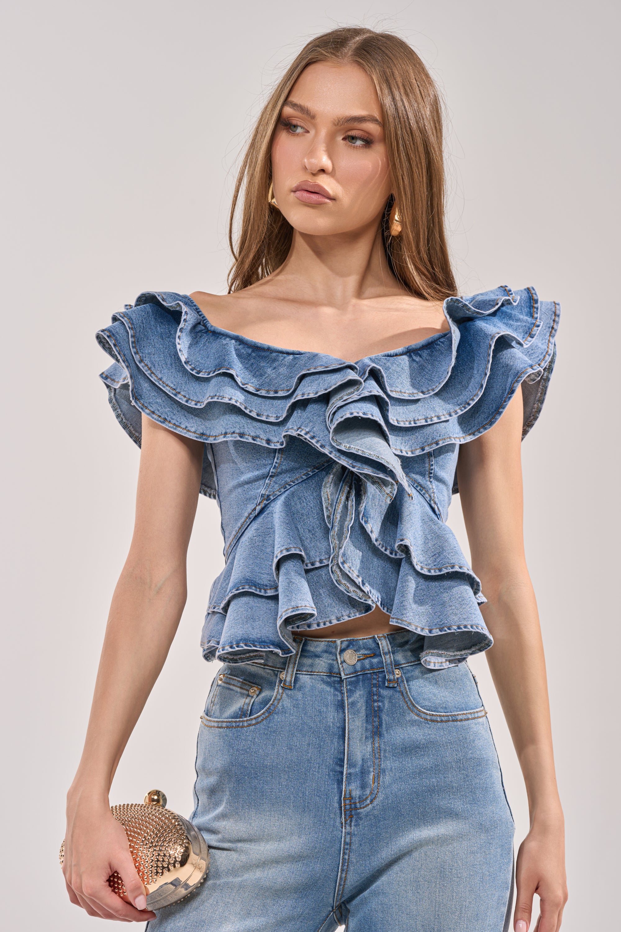 Wearing the DREAMING BIG DENIM RUFFLE TOP and high-waisted jeans, a woman with long straight hair holds a gold clutch and poses confidently against a plain light background. 1