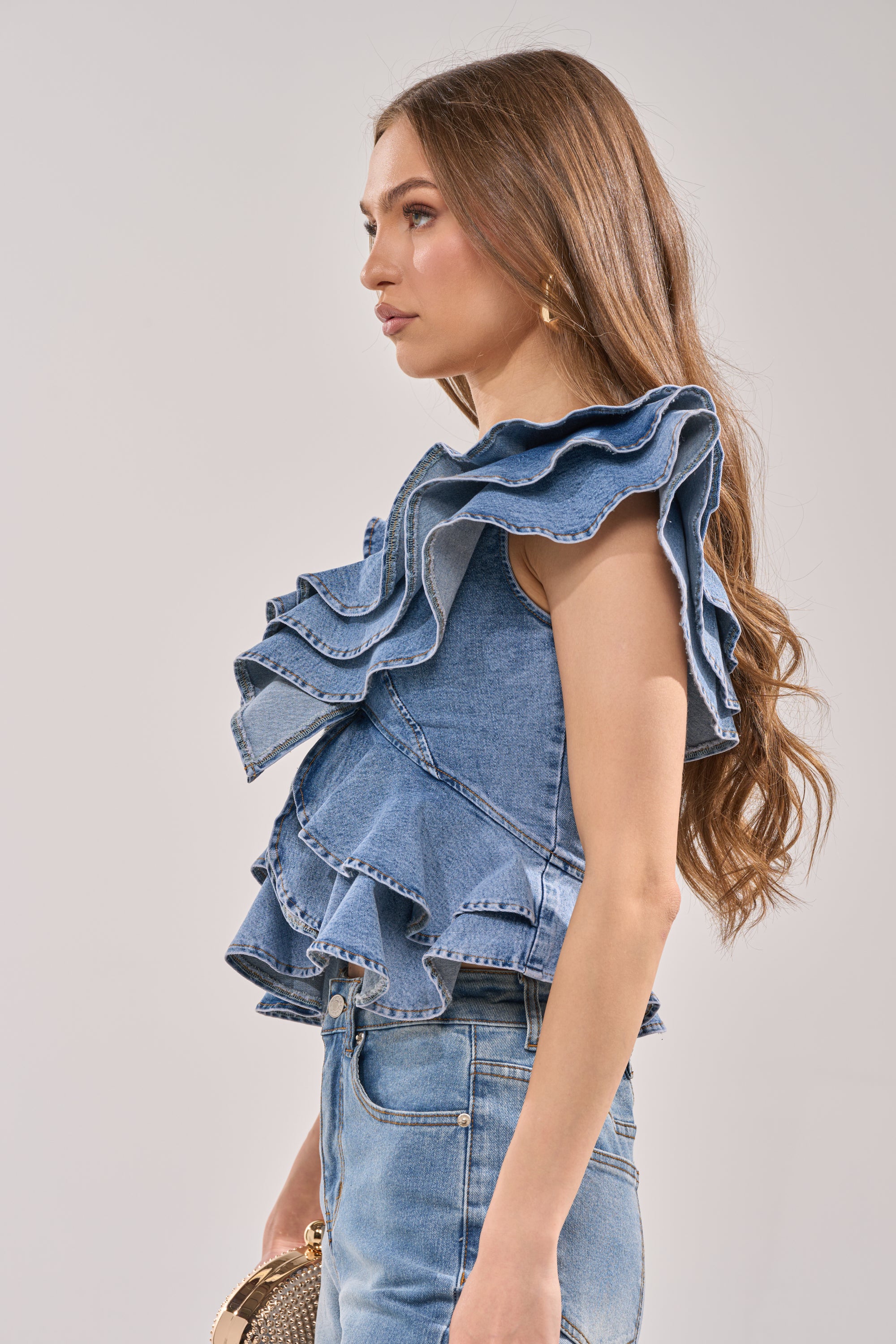 A woman with long brown hair wears the DREAMING BIG DENIM RUFFLE TOP and high-waisted denim jeans. She stands sideways against a plain light background, holding a small round handbag. 2