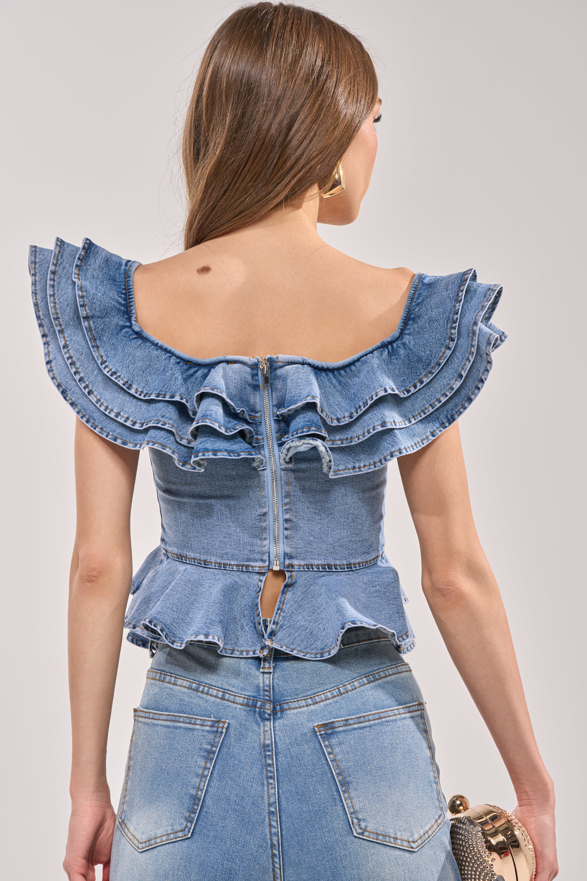 A woman with long brown hair is seen from behind, wearing the DREAMING BIG DENIM RUFFLE TOP—an off-the-shoulder peplum style with ruffled sleeves and a back zipper. She pairs it with high-waisted jeans and holds a small round purse in her right hand. 3