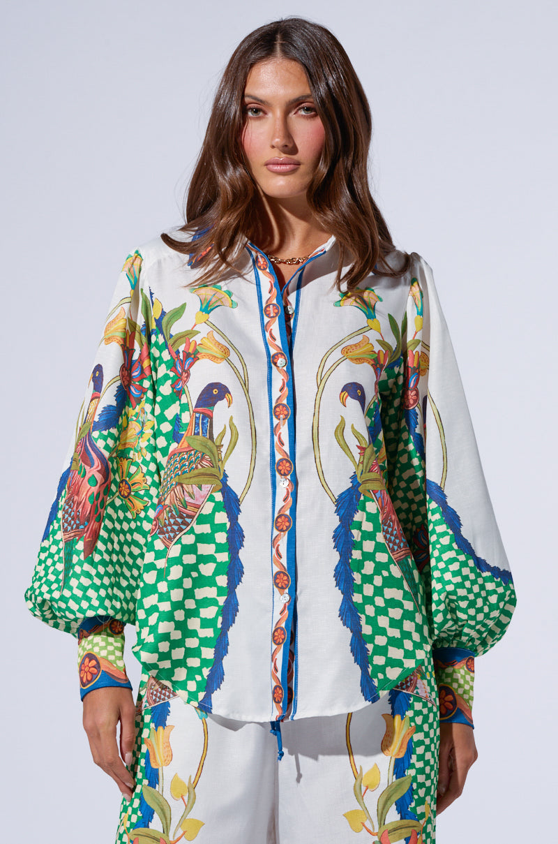 A woman with long brunette hair is wearing the WALK THIS WAY PRINTED TOP and matching pants from AKIRA Label, showcasing a vibrant pattern of peacocks, flowers, and geometric shapes in greens, blues, and oranges. The background is plain light gray.