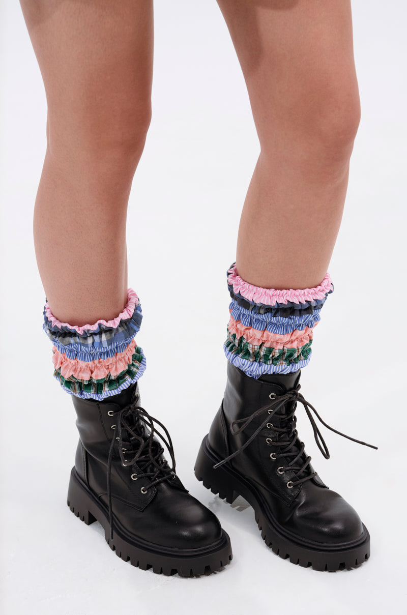 A person stands on a white background wearing the AZALEA WANG LUBNA BLACK FABRIC DETAIL BOOTIE with chunky rubber soles and colorful ruffled leg warmers in pink, blue, green, and purple. Only their legs and feet are visible.