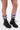 A person stands on a white background wearing the AZALEA WANG LUBNA BLACK FABRIC DETAIL BOOTIE with chunky rubber soles and colorful ruffled leg warmers in pink, blue, green, and purple. Only their legs and feet are visible.