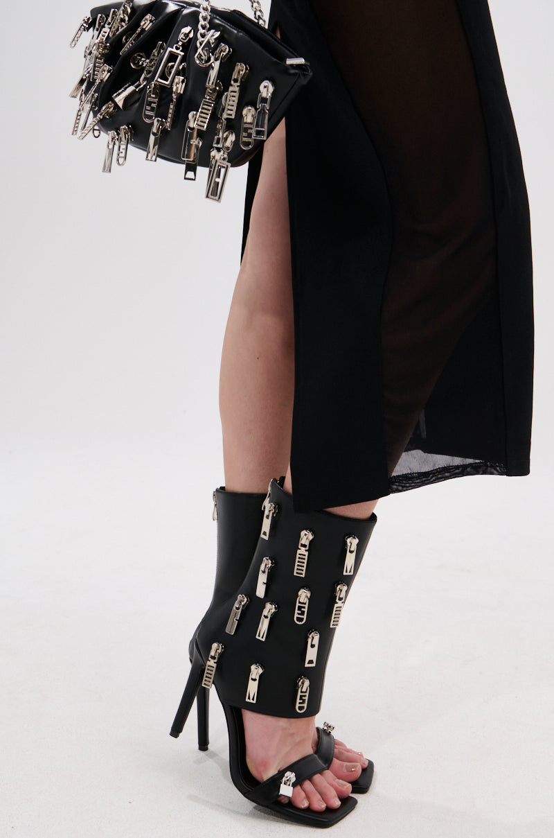Wearing the AZALEA WANG LUCAS BLACK ZIPPER DETAIL SANDAL BOOTIE with a sheer black skirt, and carrying a matching black handbag adorned with zippers.