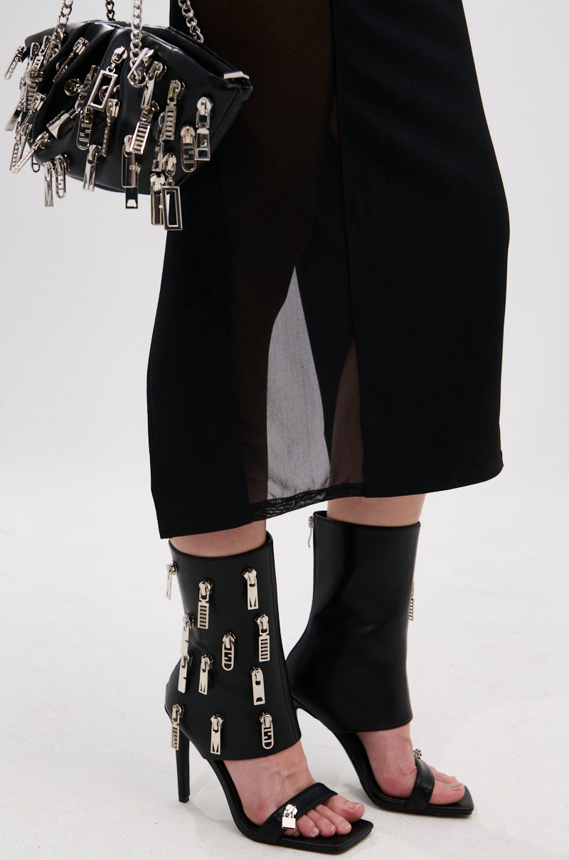 A person stands against a white background wearing the AZALEA WANG LUCAS BLACK ZIPPER DETAIL SANDAL BOOTIE and carrying a matching handbag, both in black faux leather with silver zipper pulls for a bold, stylish look.