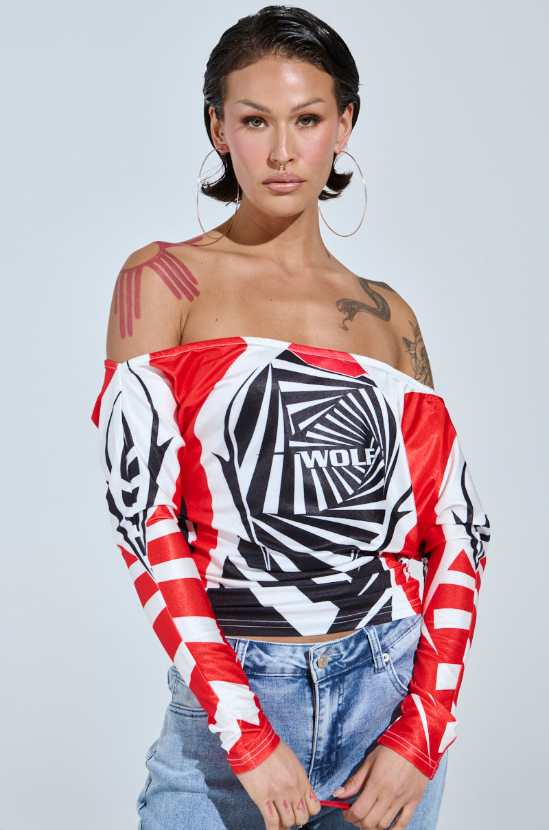 A person with large hoop earrings and slicked-back hair exudes a streetwear vibe in a FOX RACING OFF THE SHOULDER LIGHT WEIGHT TEE featuring bold red, black, and white geometric patterns, paired with blue jeans. Tattoos adorn their shoulder and wrist.