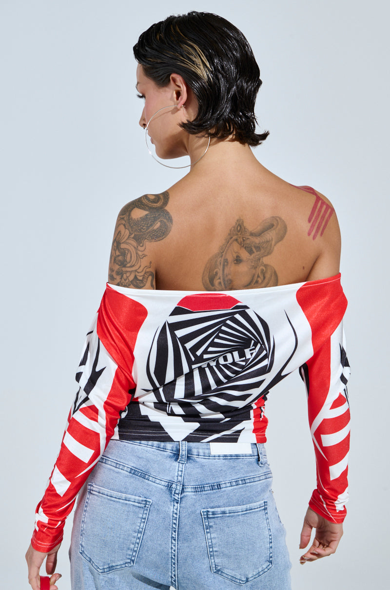 Someone with short, dark hair gives off a streetwear vibe in a FOX RACING OFF THE SHOULDER LIGHT WEIGHT TEE featuring red, black, and white abstract patterns. Tattoos peek from their upper back and shoulder, while light blue jeans complete the ensemble against a plain, light backdrop.