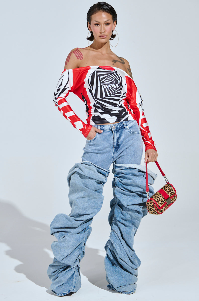 Confidently styled, a person dons the FOX RACING OFF THE SHOULDER LIGHTWEIGHT TEE in red, black, and white patterns with oversized blue ruched jeans. They carry a small red and leopard print handbag, while large hoop earrings complete the streetwear vibe of this fashion-forward ensemble.