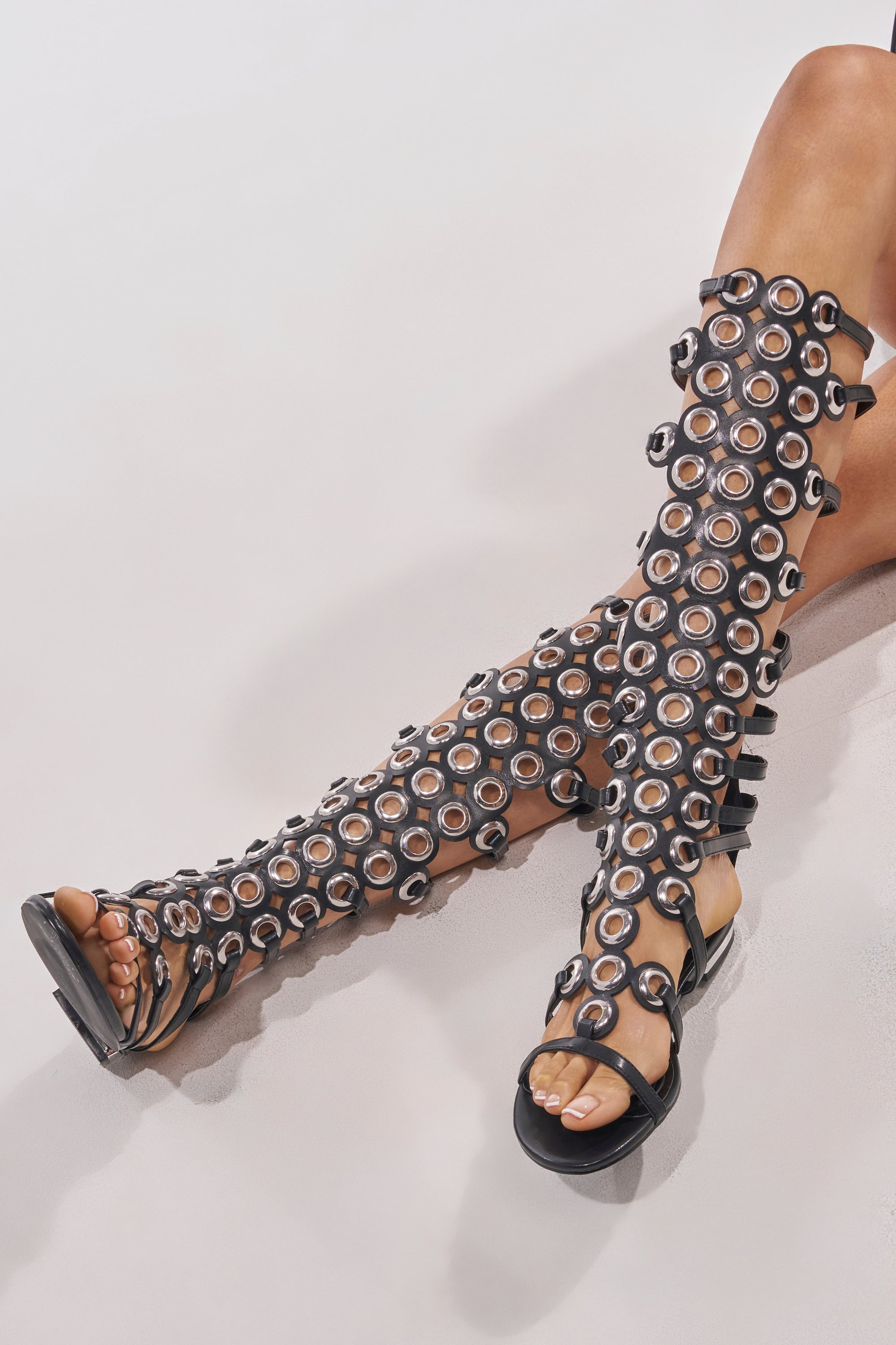 Seated with legs extended and crossed, a person wears AZALEA WANG LUCE BLACK GROMMET DETAIL FLAT GLADIATOR SANDALS—knee-high faux leather featuring circular cutouts and metallic ring accents—on a white surface. 2