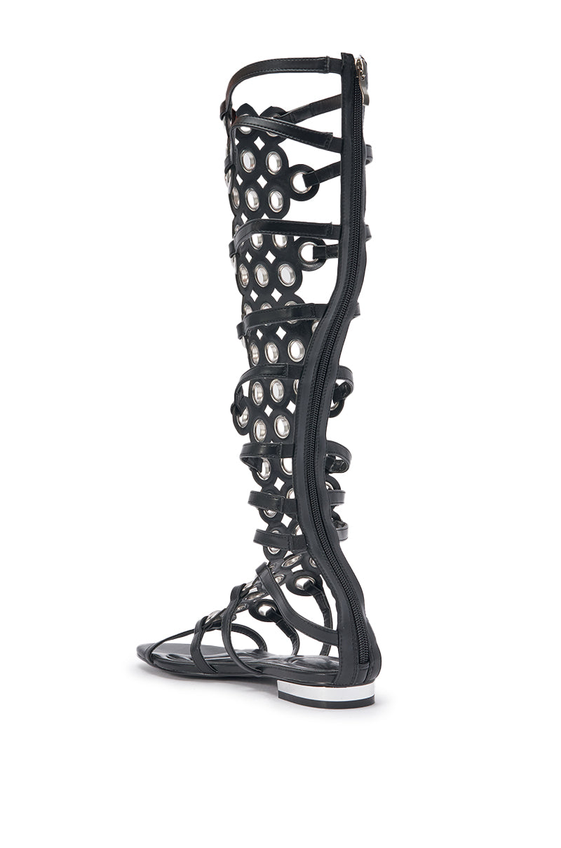 The AZALEA WANG LUCE BLACK GROMMET DETAIL FLAT GLADIATOR SANDAL features black faux leather, circular cut-outs, knee-high fit, silver grommet accents, and a side zipper for easy wear. 5