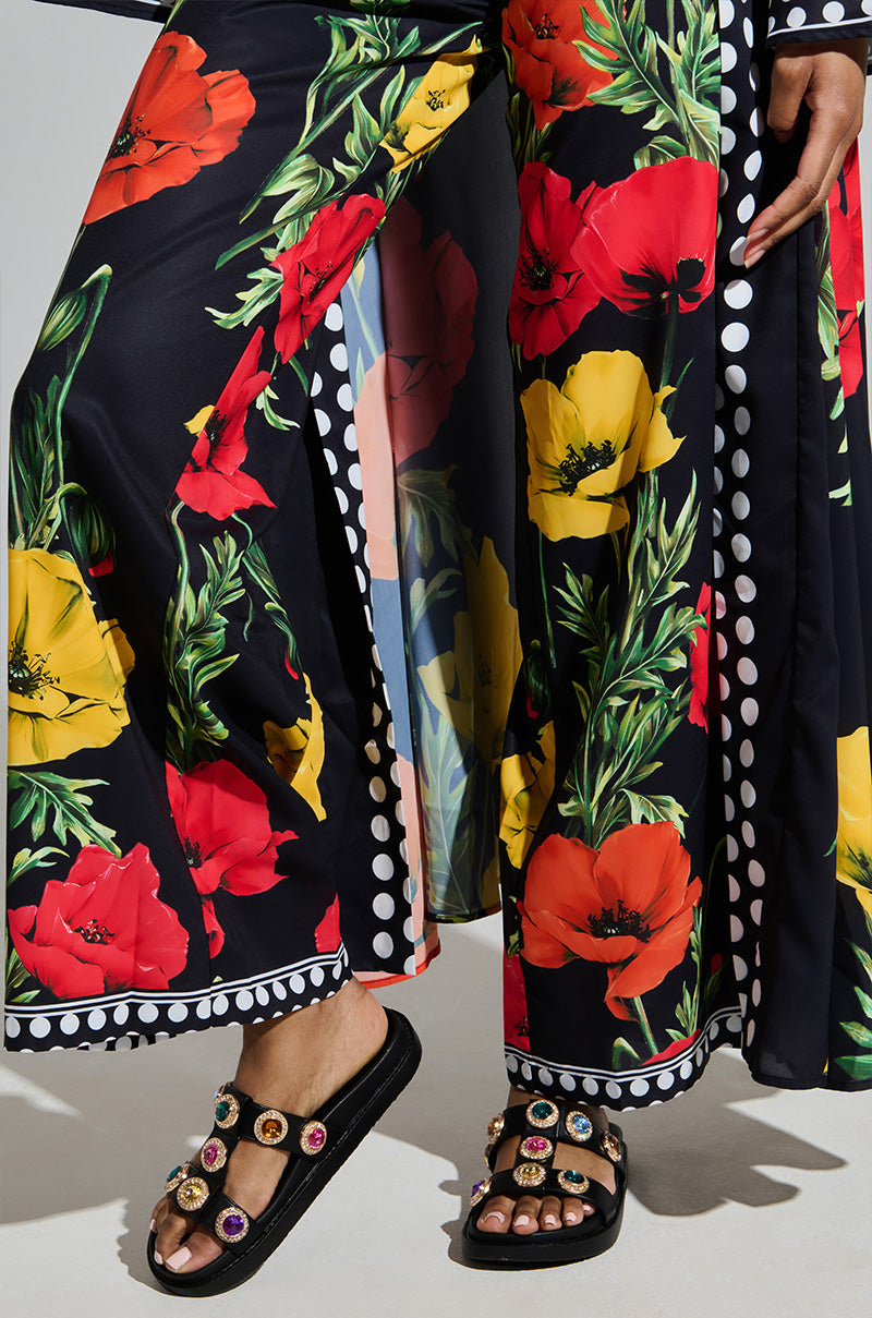 Someone is wearing vibrant wide-leg pants featuring red and yellow poppies with green leaves, bordered by black and white polka dots. They're pairing them with AZALEA WANG LUCIENA BLACK RHINESTONE SANDAL, which has dazzling gemstone details.
