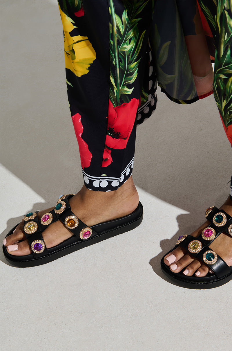 A person wearing AZALEA WANG LUCIENA BLACK RHINESTONE SANDALS adorned with colorful jewels. Their pants flaunt a vibrant floral design in red, yellow, and green on a black background. Natural sunlight beautifully illuminates the scene.