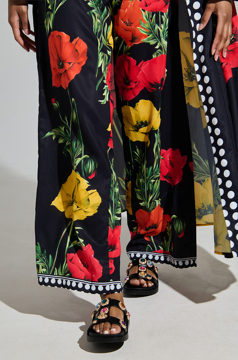 A person wearing black pants with bold red and yellow floral prints pairs them perfectly with the AZALEA WANG LUCIENA BLACK RHINESTONE SANDALS, adorned with colorful embellishments. The bottom hem flaunts a chic black and white polka dot pattern.
