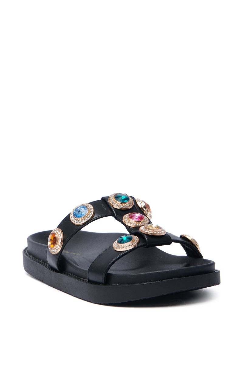 The AZALEA WANG LUCIENA Black Rhinestone Sandal features a flat sole and wide criss-cross black vegan leather straps with colorful gemstones set in gold-tone settings, adding a decorative touch.
