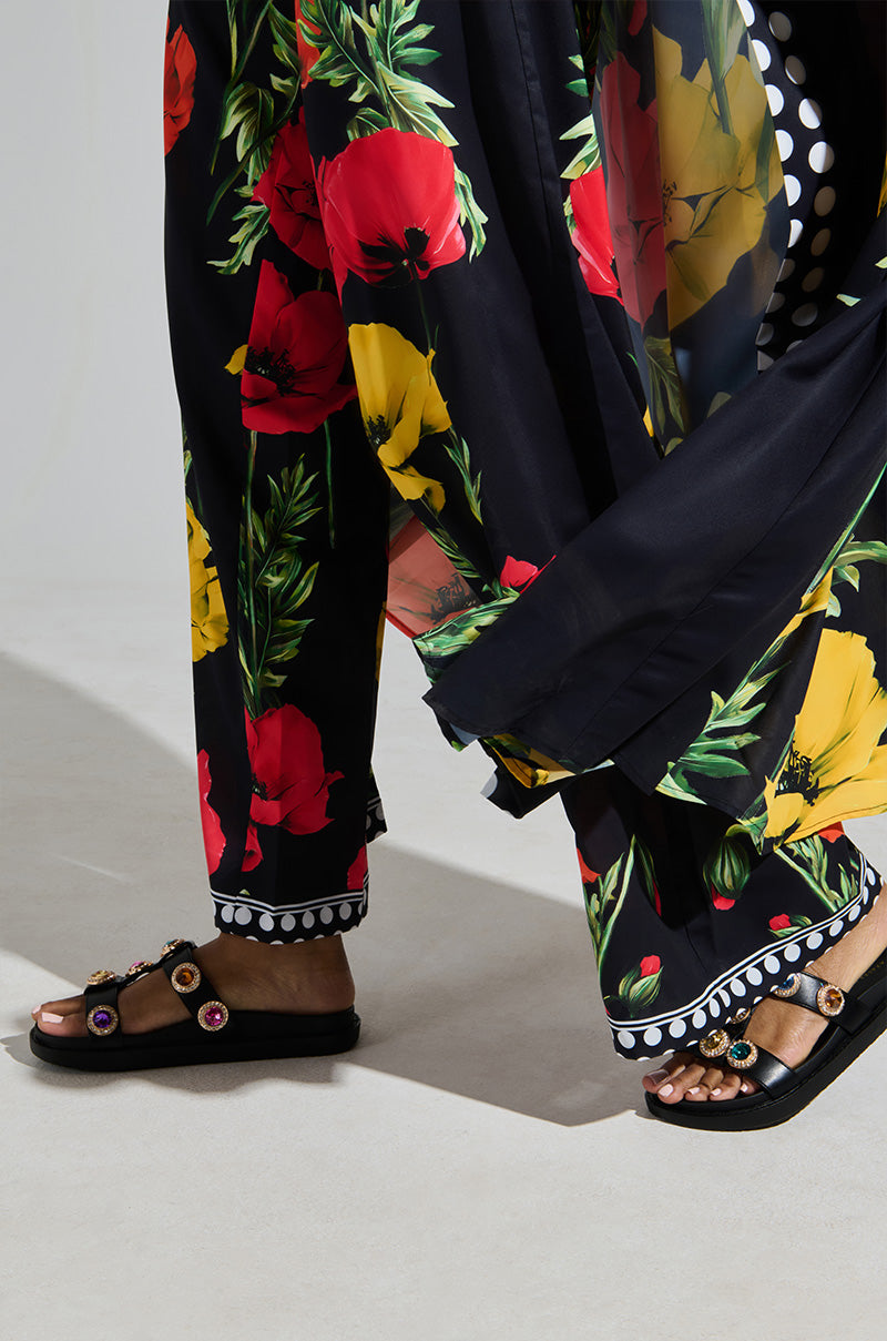 Confidently walking in a black garment with vibrant floral prints, the person proudly showcases their AZALEA WANG LUCIENA BLACK RHINESTONE SANDAL. The chic look is completed by the hem's stylish black and white polka dot trim.
