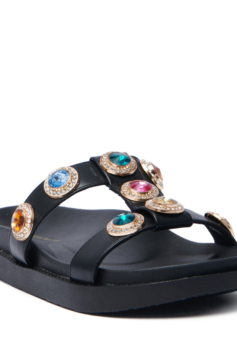 The AZALEA WANG LUCIENA BLACK RHINESTONE SANDAL features black vegan leather crisscross straps adorned with large, vibrant blue, green, pink, and yellow gemstones set in gold-colored settings. Its flat sole enhances the eye-catching design.