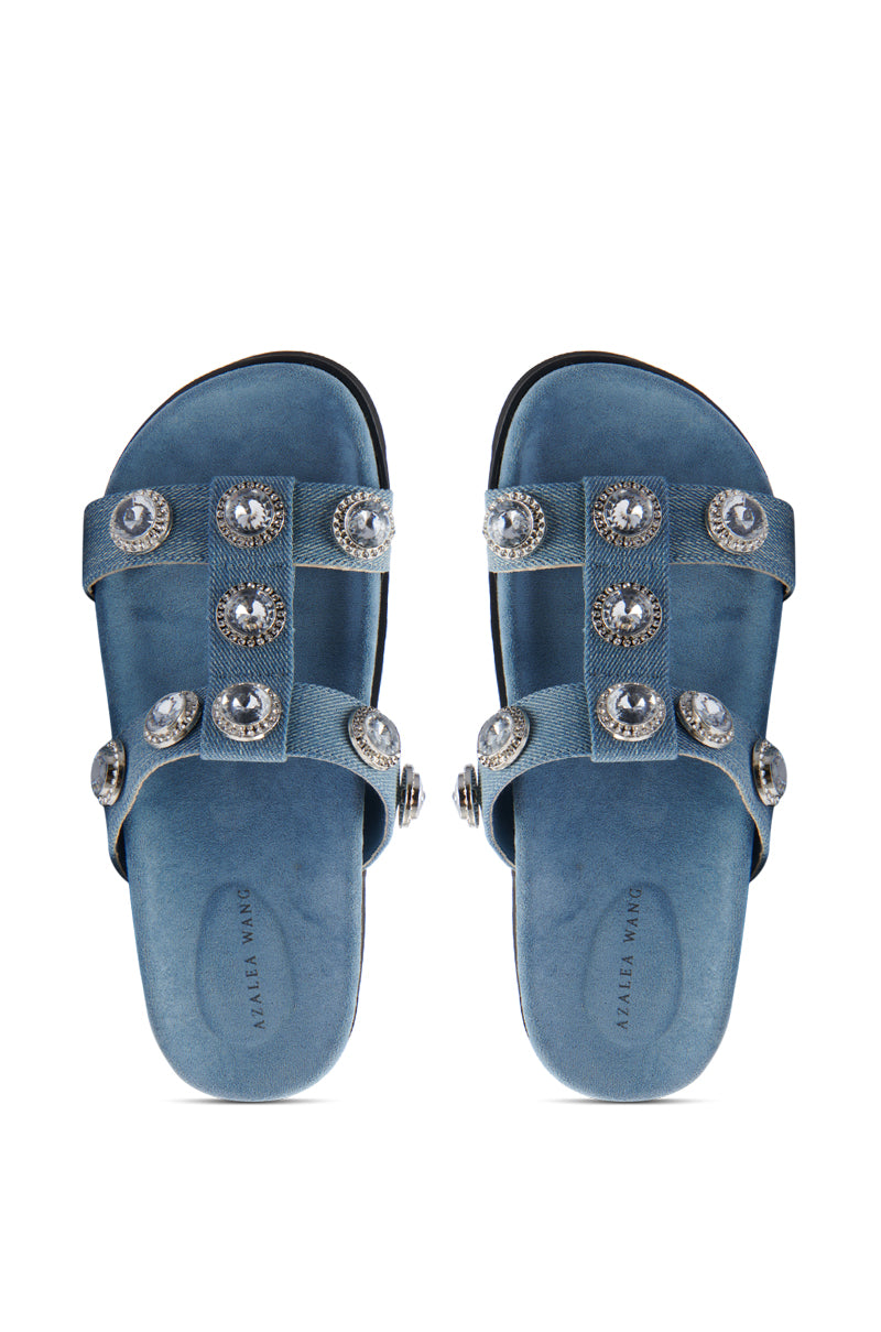 The AZALEA WANG LUCINEA DENIM RHINESTONE SANDAL features blue slip-on denim sandals with two straps adorned with large silver studs and a suede insole embossed with "TASSEL WANG," adding casual flair to any outfit.
