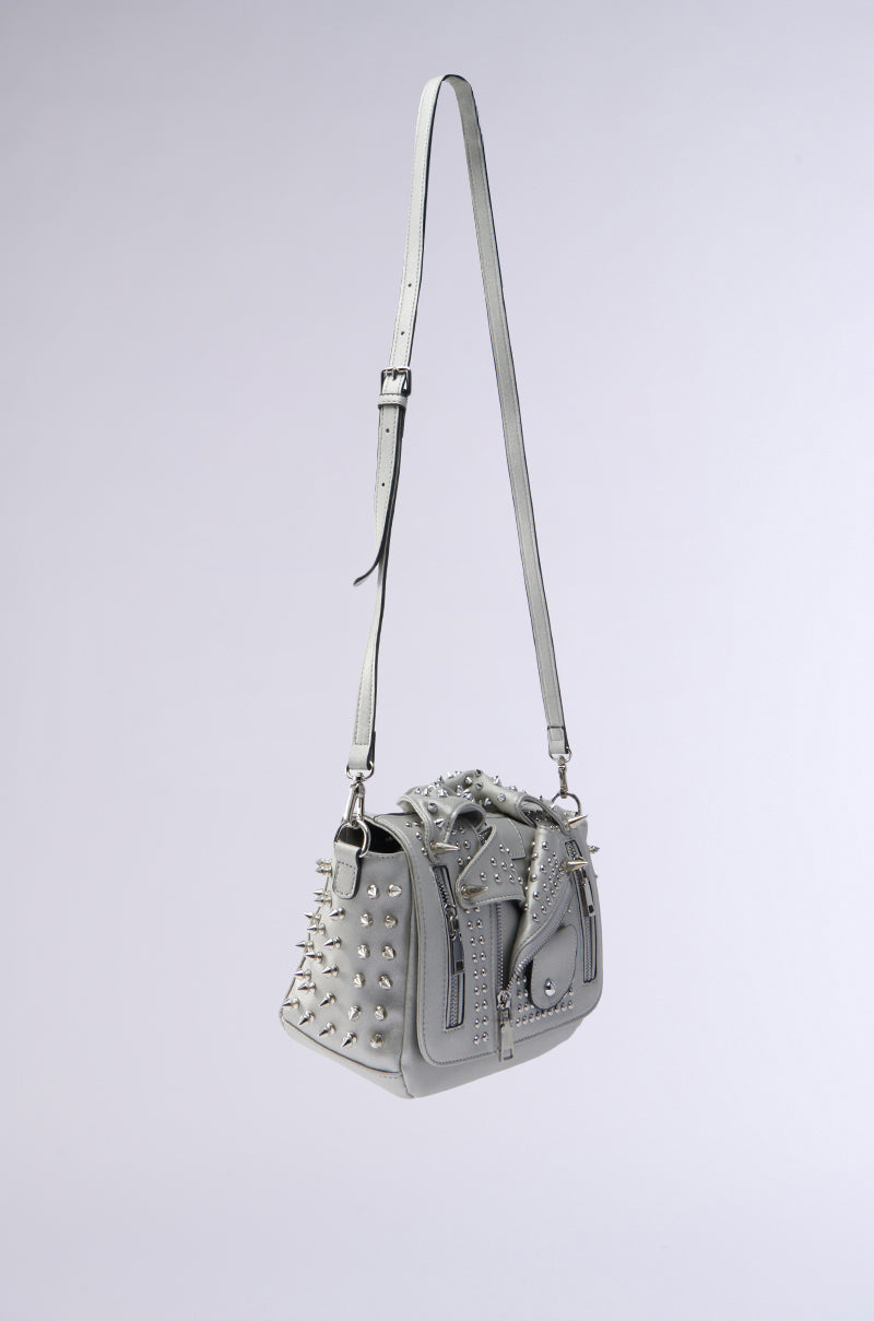 The MOTO OF YOUR DREAMS PURSE is a gray faux leather bag with silver studs, decorative zippers, and a long adjustable strap. It has a structured shape with unique metal embellishments, echoing the look of a moto jacket.